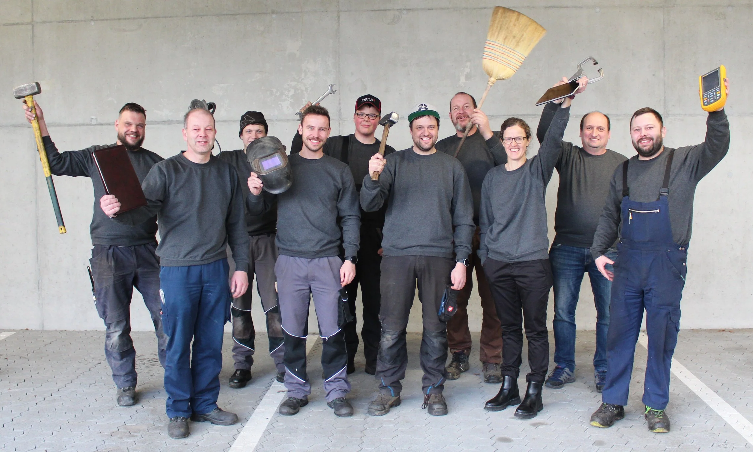 Gruppe von zehn Menschen, die als Handwerker gekleidet sind, stehen vor einer grauen Betonwand und posieren für das Foto, einige halten Werkzeuge in den Händen.