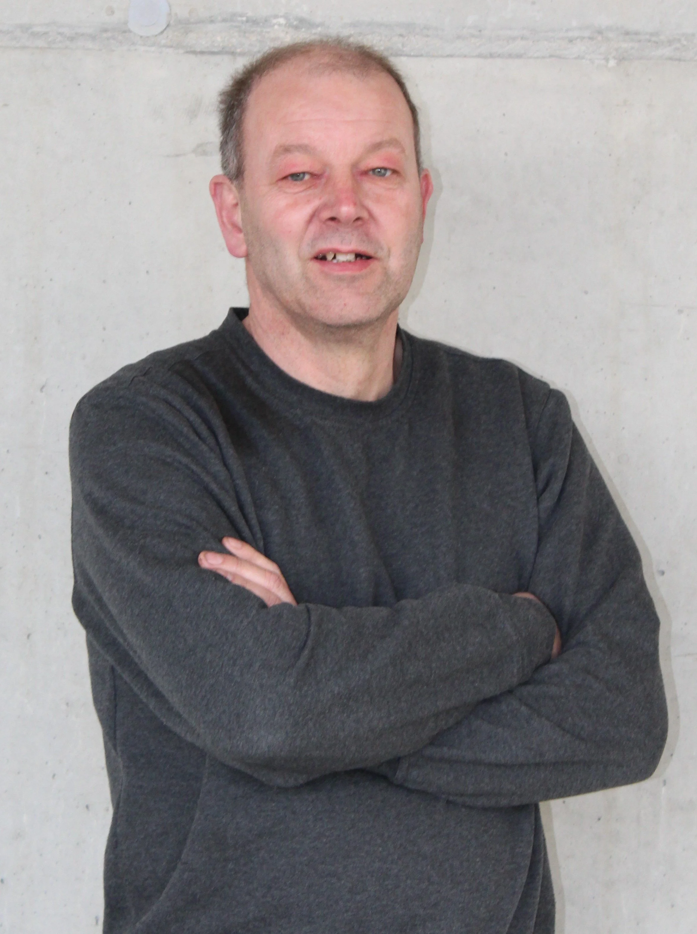 Ein Mann mit kurzgeschnittenem Haar, trägt ein dunkles Sweatshirt, steht vor einer glatten, hellgrauen Wand und hat die Arme verschränkt.