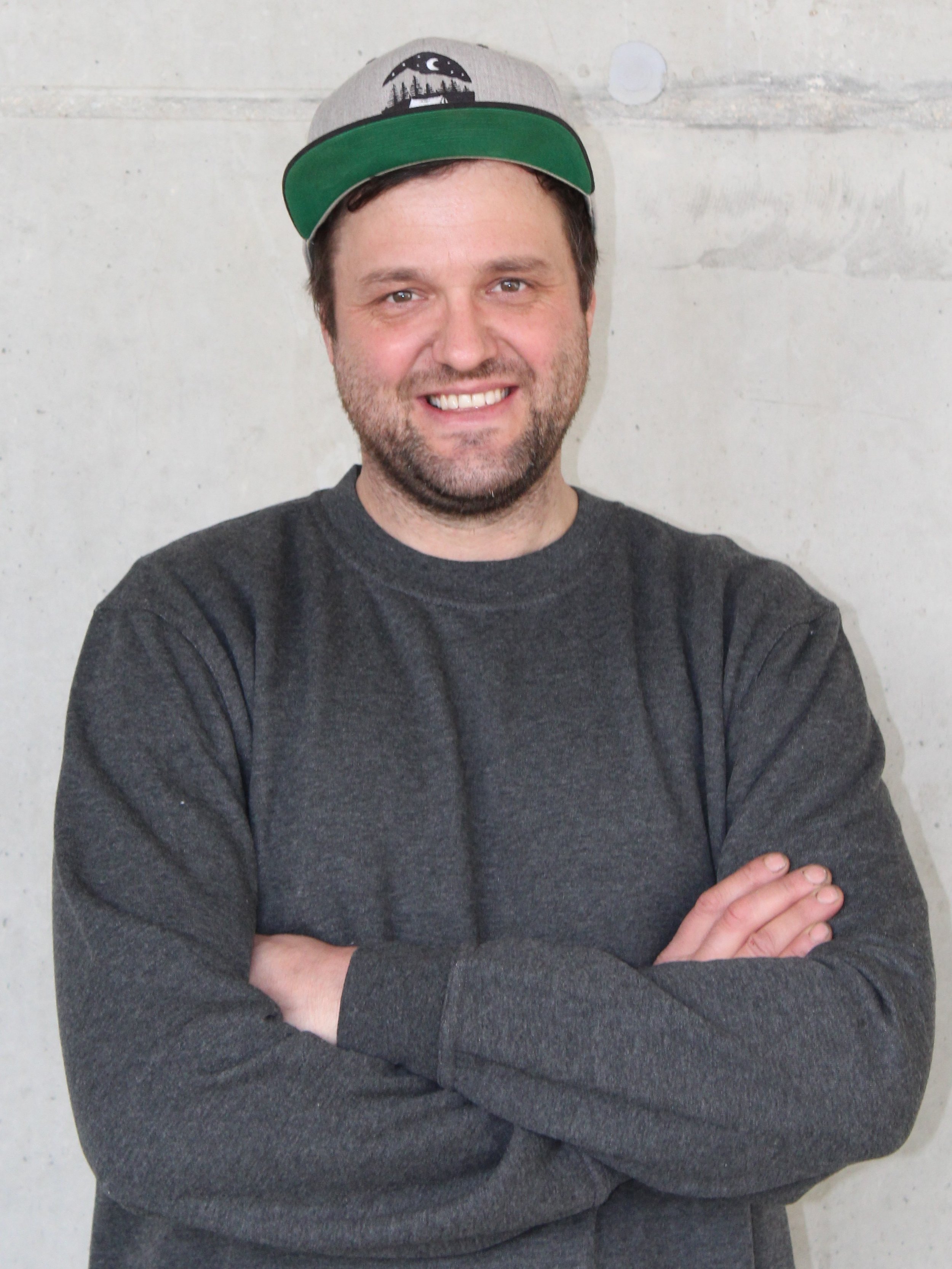 Ein Mann mit dunklem Haar, Bart und schwarzen Augen steht vor einer hellen Wand, trägt ein graues sweatshirt und einen grau-grünen Baseballcap mit einem schwarzen und weißen Design.
