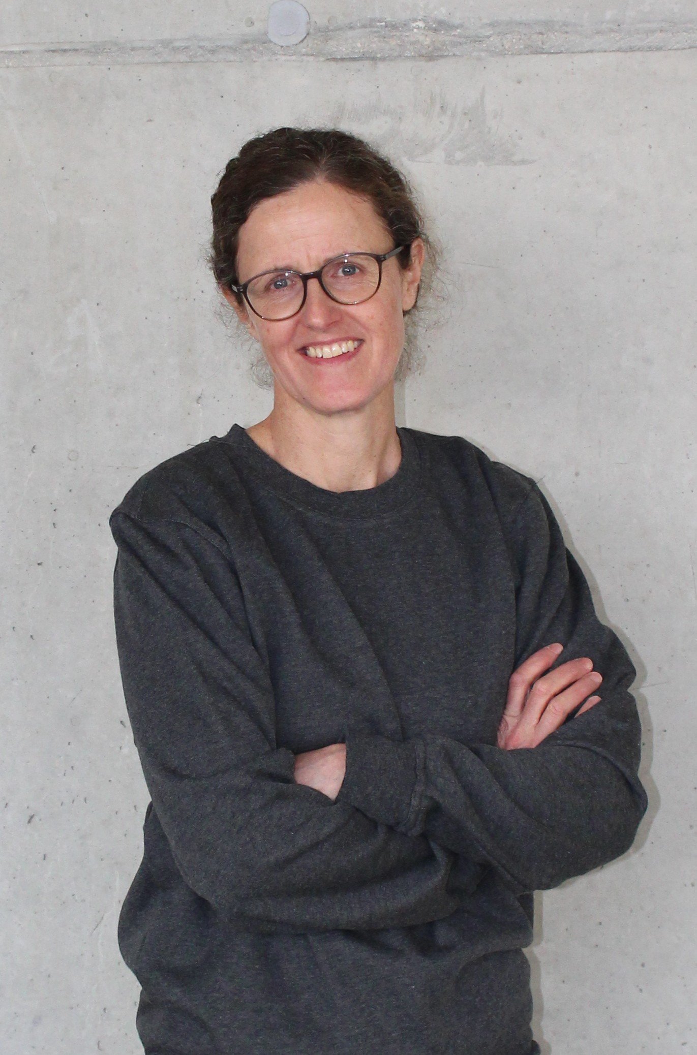 Frau mit Brille und schwarzen Pullover, Arme verschränkt, vor einer Betonwand.
