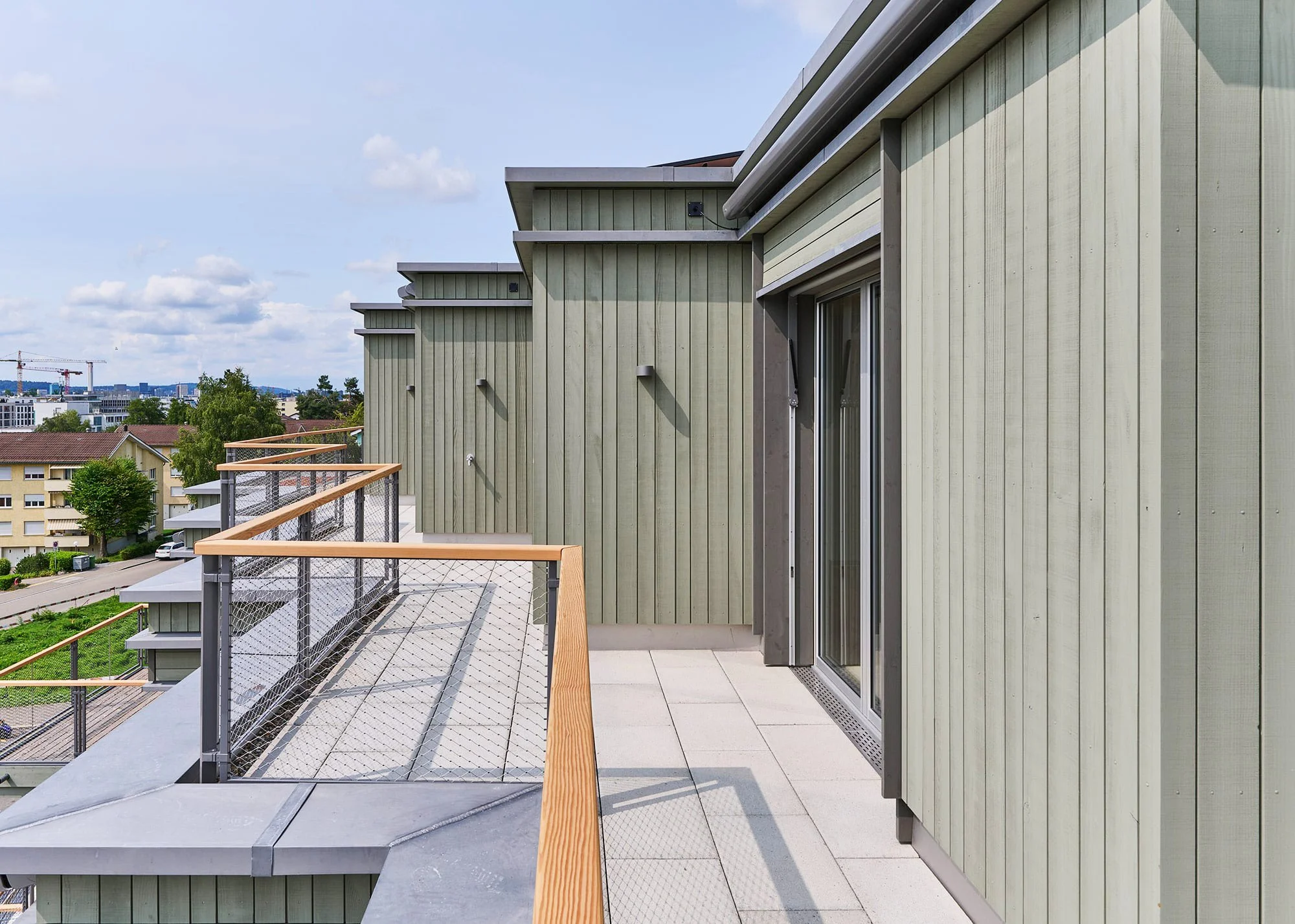 Balkonansicht eines modernen Gebäudes mit grünem Holzausbau, Glas-Schiebetüren, Holz- und Metallbrüstungen, in einer urbanen Umgebung bei sonnigem Wetter.