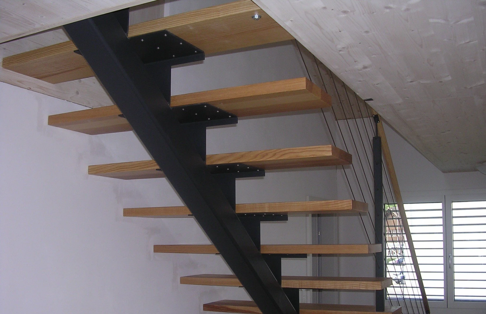 Holztreppe mit schwarzen Metallhalterungen im Innenraum, darunter ein Fenster mit Gitter, und sichtbare Decke aus Holzpaneelen.