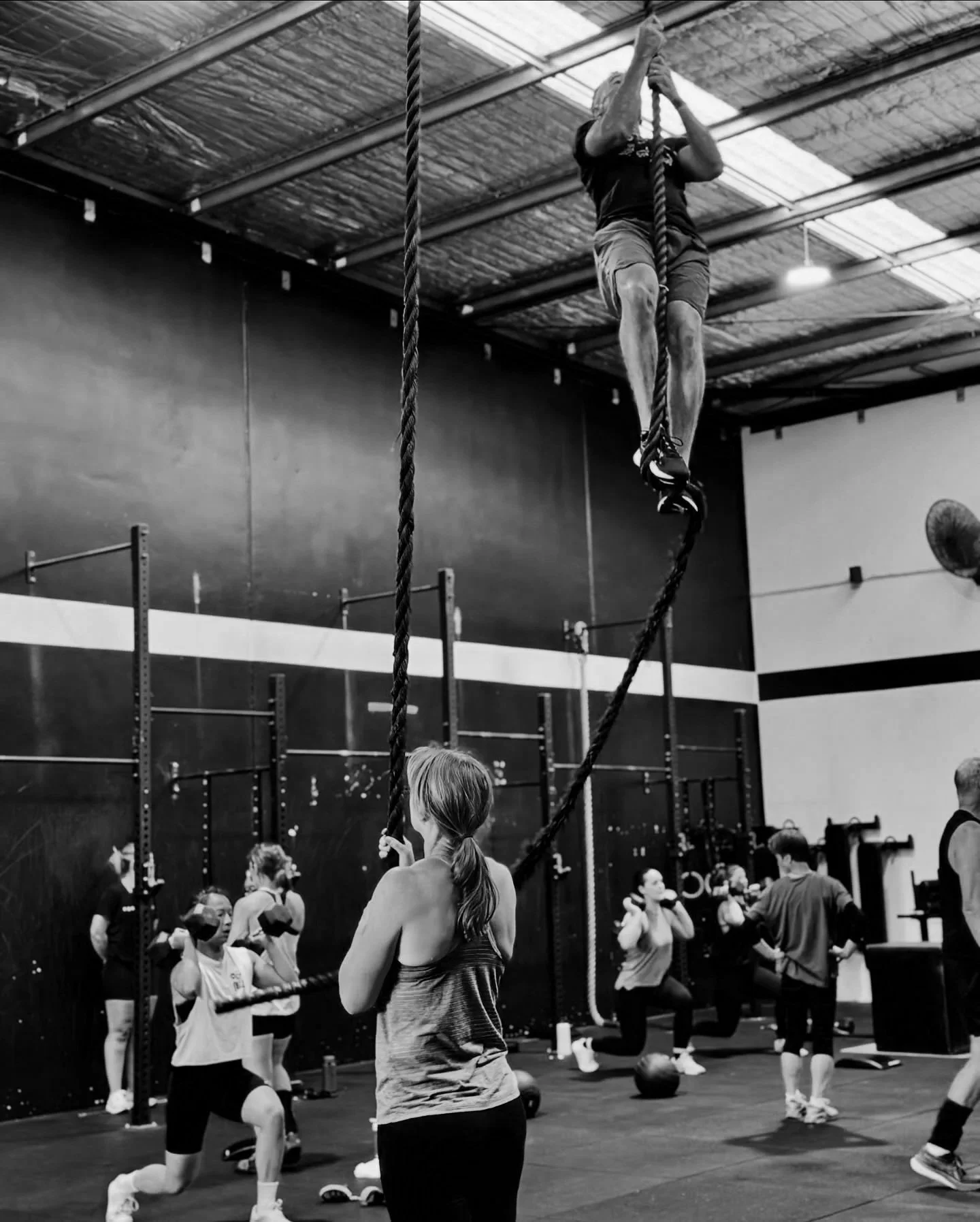 People participating in a fitness class at a gym, including a woman holding onto battle ropes, with others in the background doing exercises.