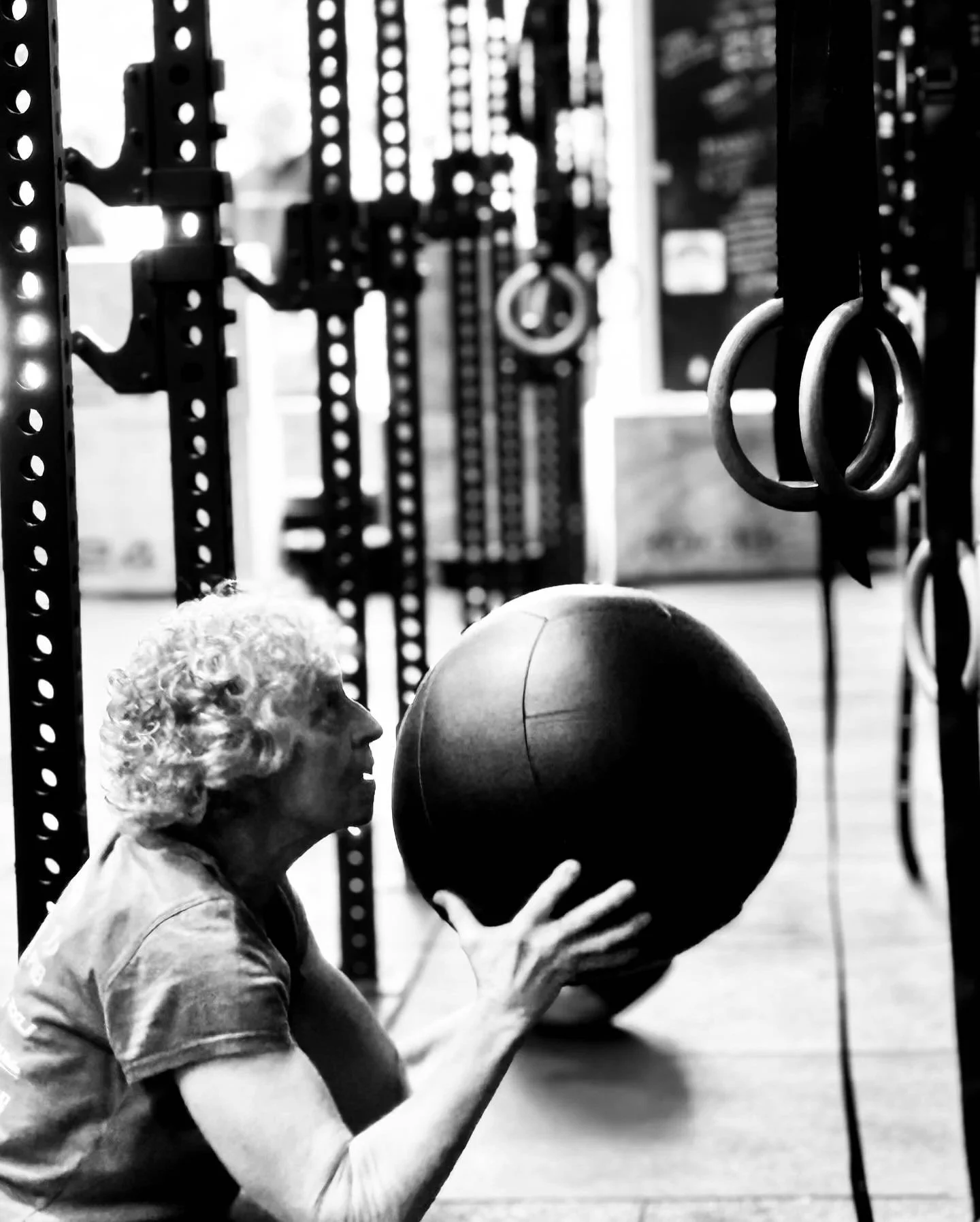 An elderly woman with curly hair is holding a medicine ball during a workout in a gym, with gym equipment visible in the background.