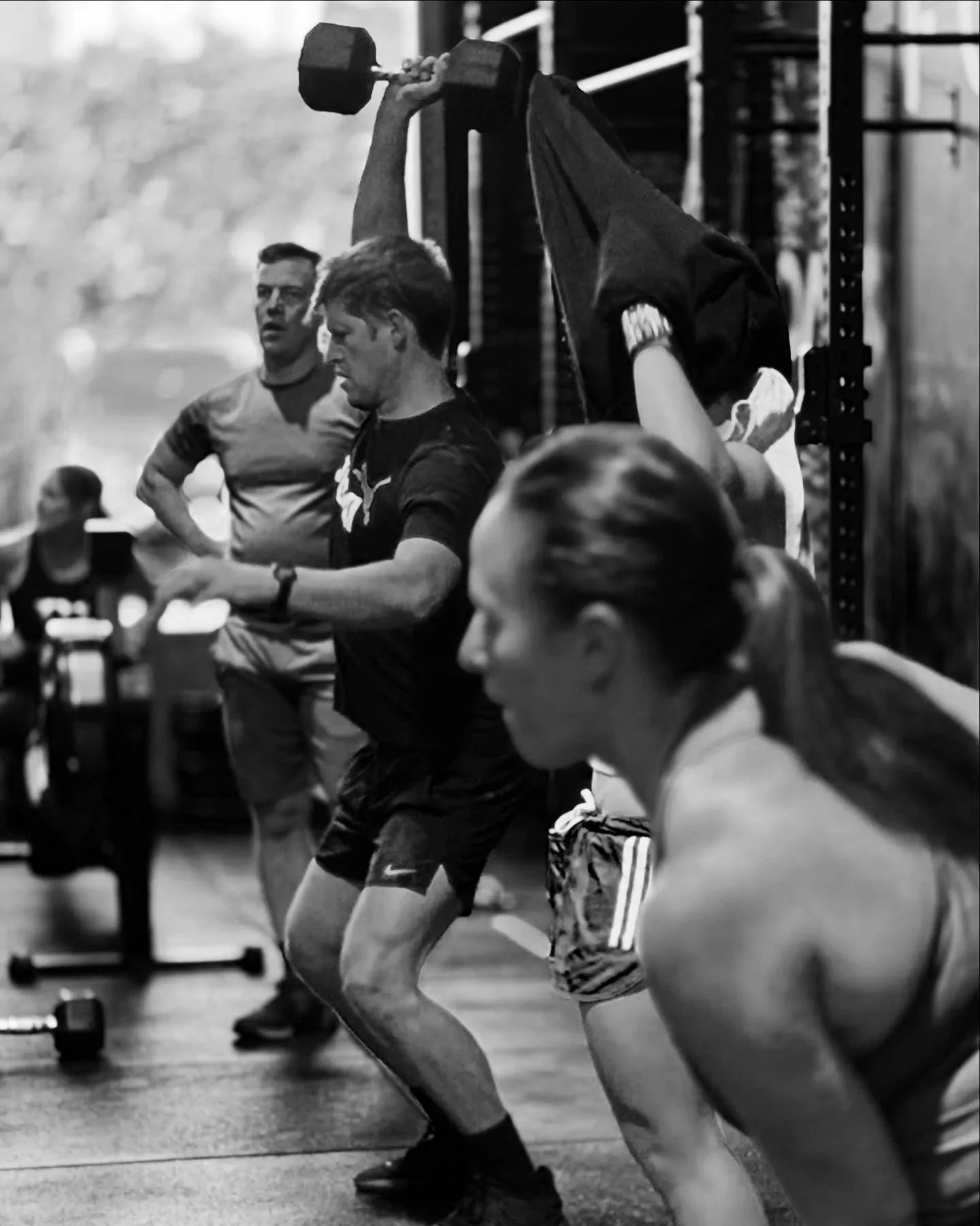 People in a gym performing a workout, with one person lifting a dumbbell and others observing or waiting their turn.