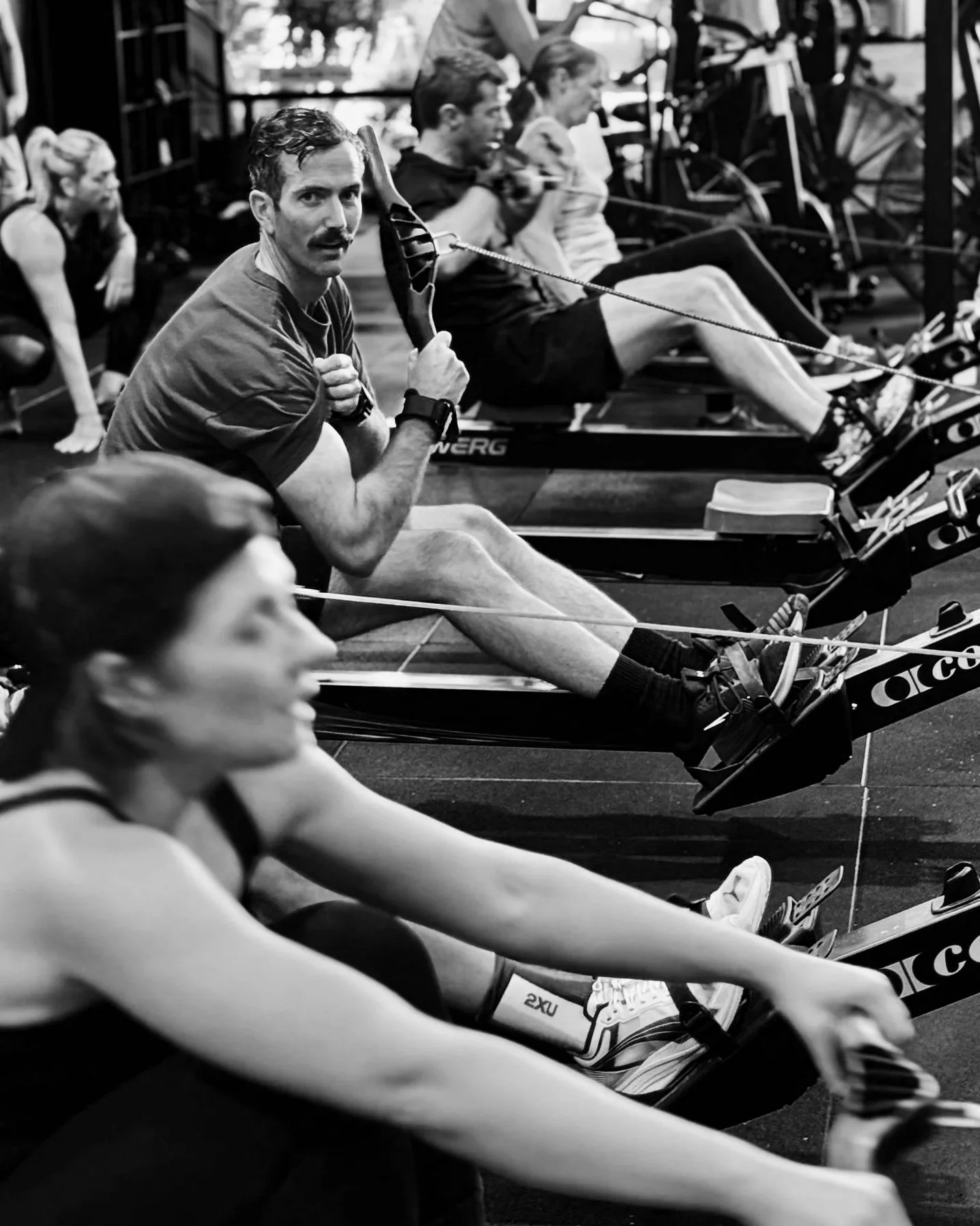 Multiple people engaging in a rowing workout at a gym, sitting on rowing machines, with one man looking towards the camera in the foreground.