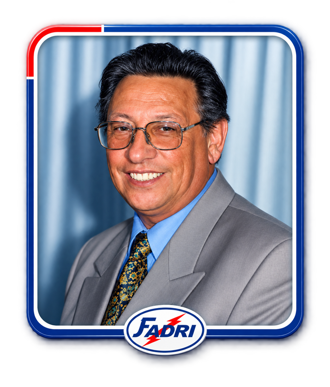 Hombre de mediana edad con gafas, cabello oscuro peinado hacia atrás, vistiendo un saco gris, camisa azul y corbata con patrón, sonriendo en un retrato profesional con fondo de cortina azul y un marco con los colores de la bandera y el logo de FADRI.