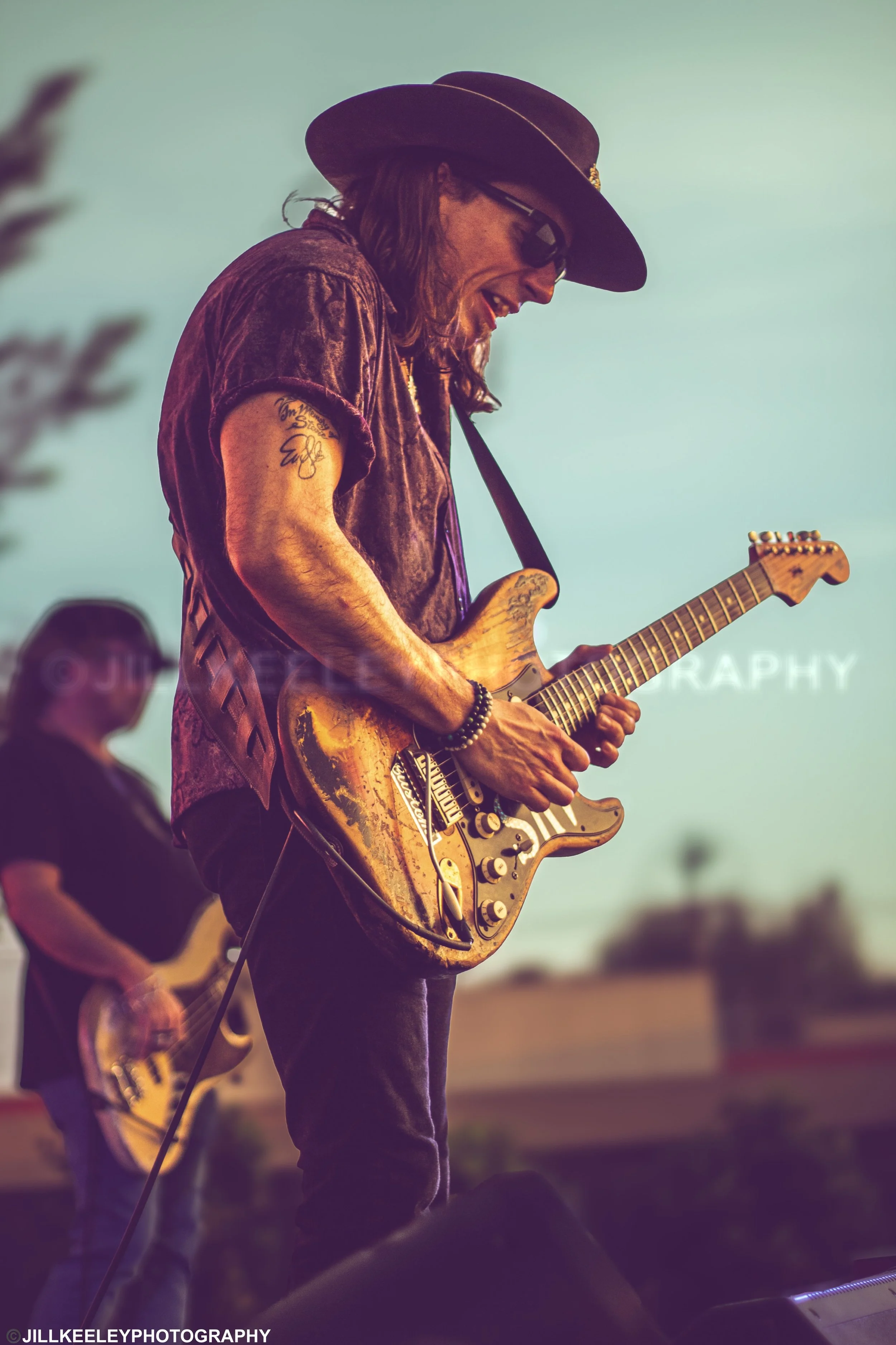 A man wearing a wide-brimmed hat, sunglasses, and a patterned shirt is playing an electric guitar during an outdoor performance at sunset, with a slightly blurred background and other band members visible.