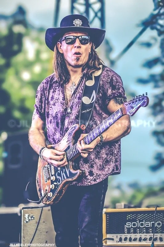 A male musician wearing a wide-brimmed hat, sunglasses, and a purple patterned shirt, playing an electric guitar during a live outdoor performance.