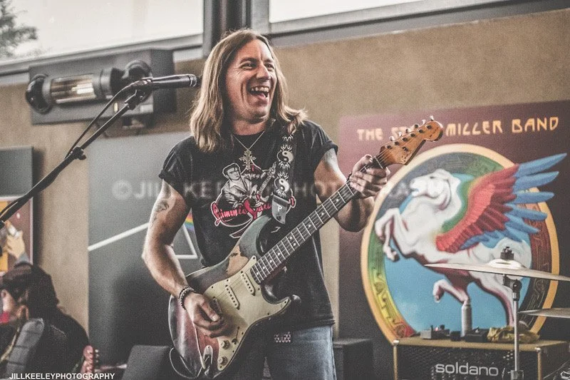 A male musician with long hair playing an electric guitar and smiling during a performance at a venue.