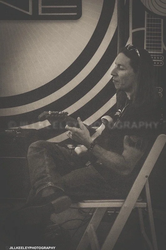 A guitarist  sitting on a chair playing an electric guitar against a decorated wall with circular patterns.