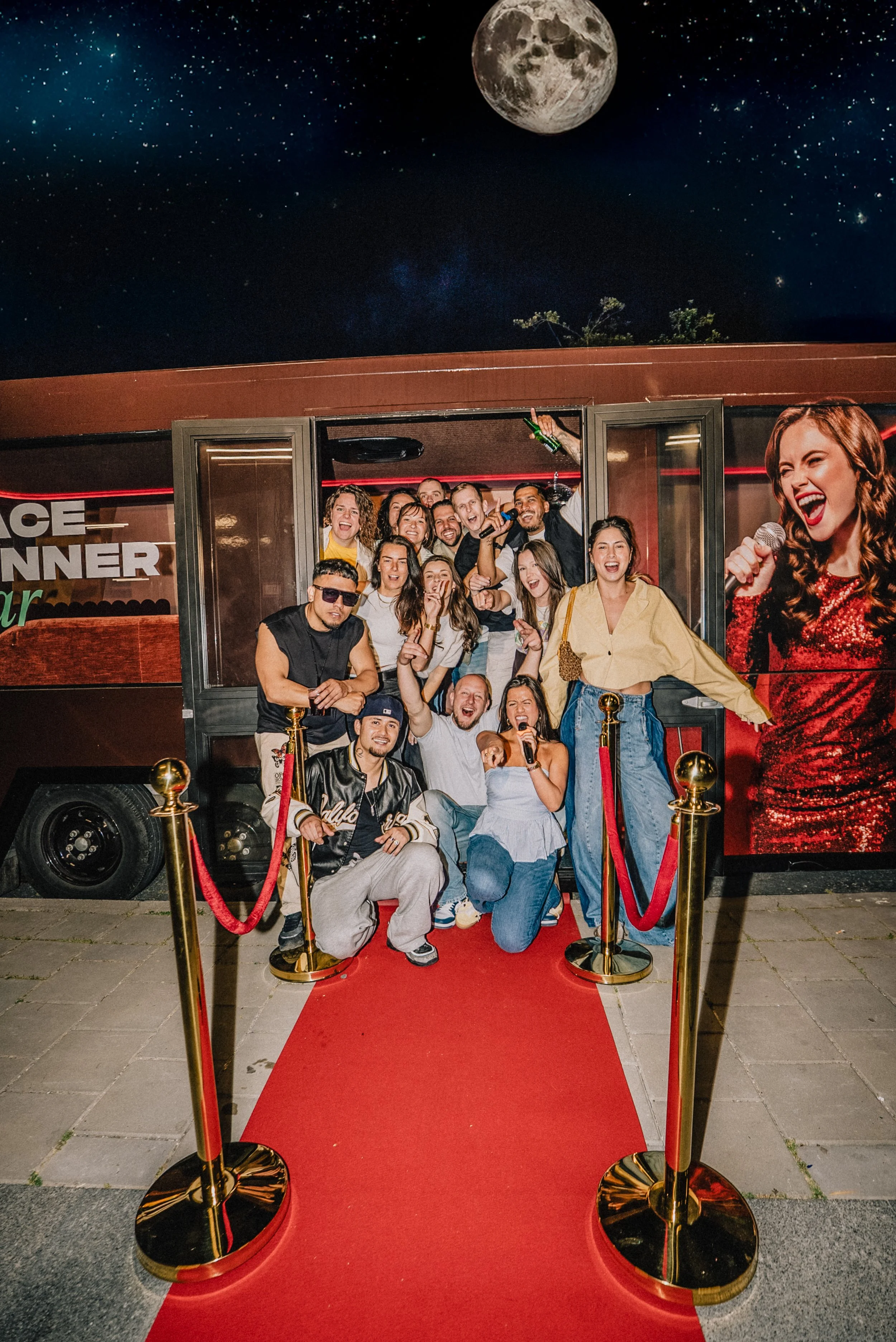 Groep mensen posed op een rode loper voor een bus met de tekst 'Face Winner' in de avond, onder een grote afbeelding van de volle maan en sterren op de achtergrond.