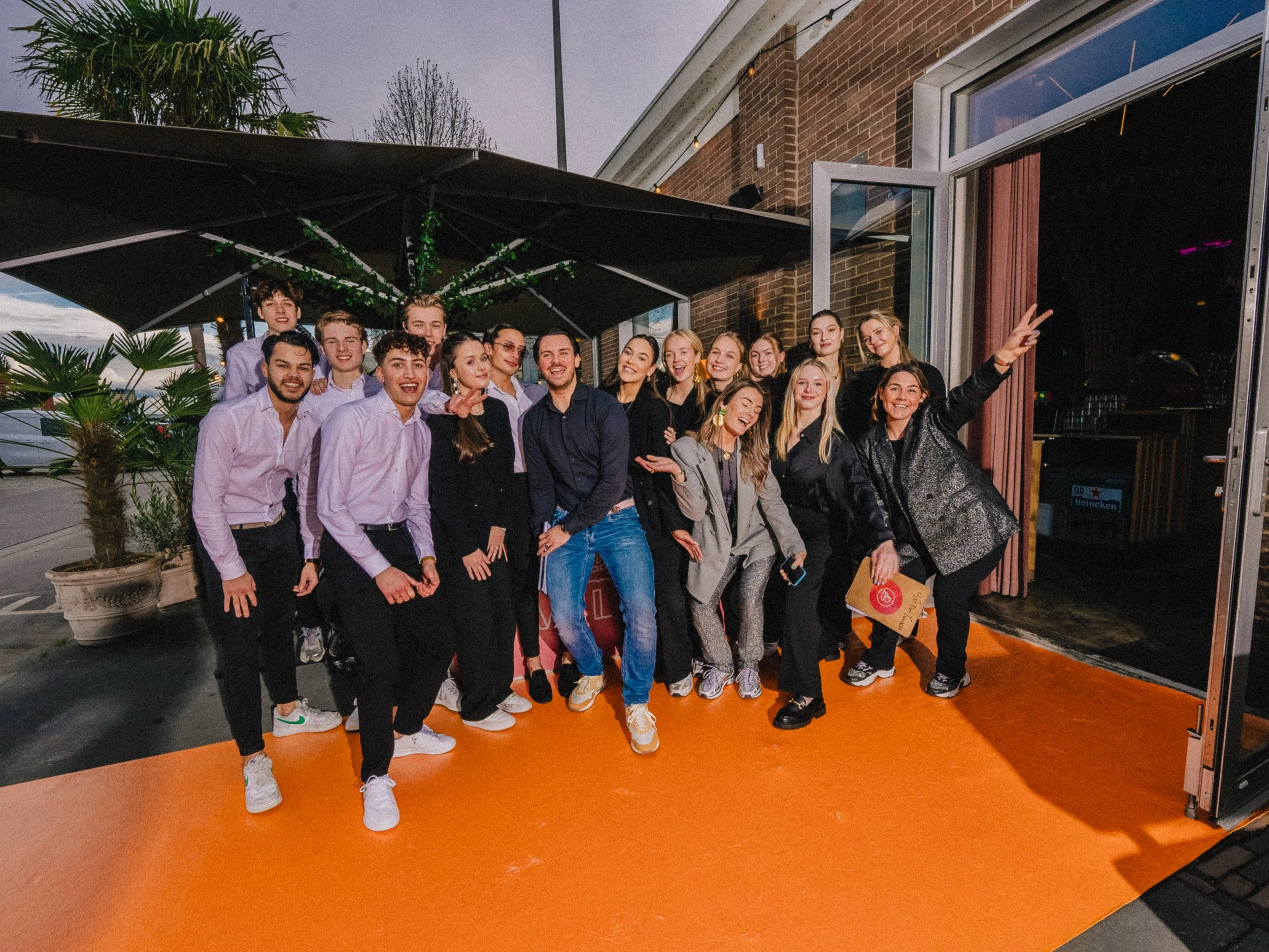 Groep jonge mensen die vrolijk poseert in een restaurant of café met een buitenbar, onder een parasol en naast een huis met gevels van baksteen, in de avond. Ze lachen en maken girafvriendelijke gebaren.
