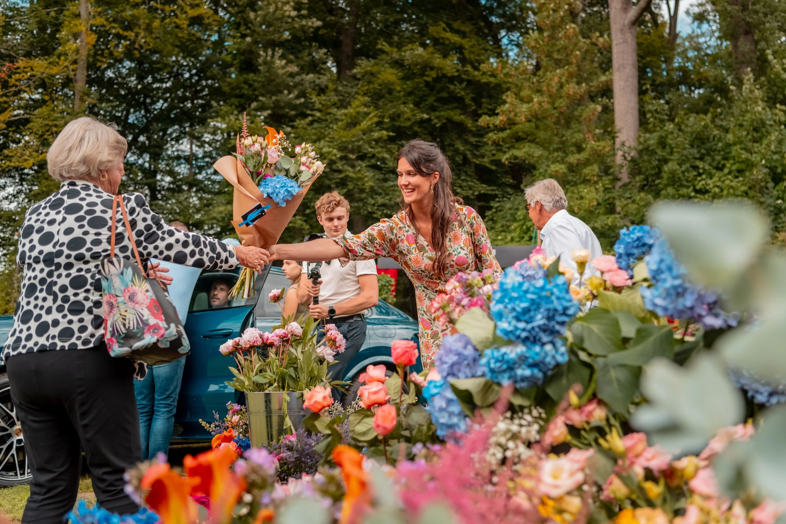 Een groep mensen op een markt met bloemen, een jonge vrouw ontvangt een bos bloemen van een oudere vrouw, terwijl anderen toekijken, en de setting is in een bosrijke omgeving.