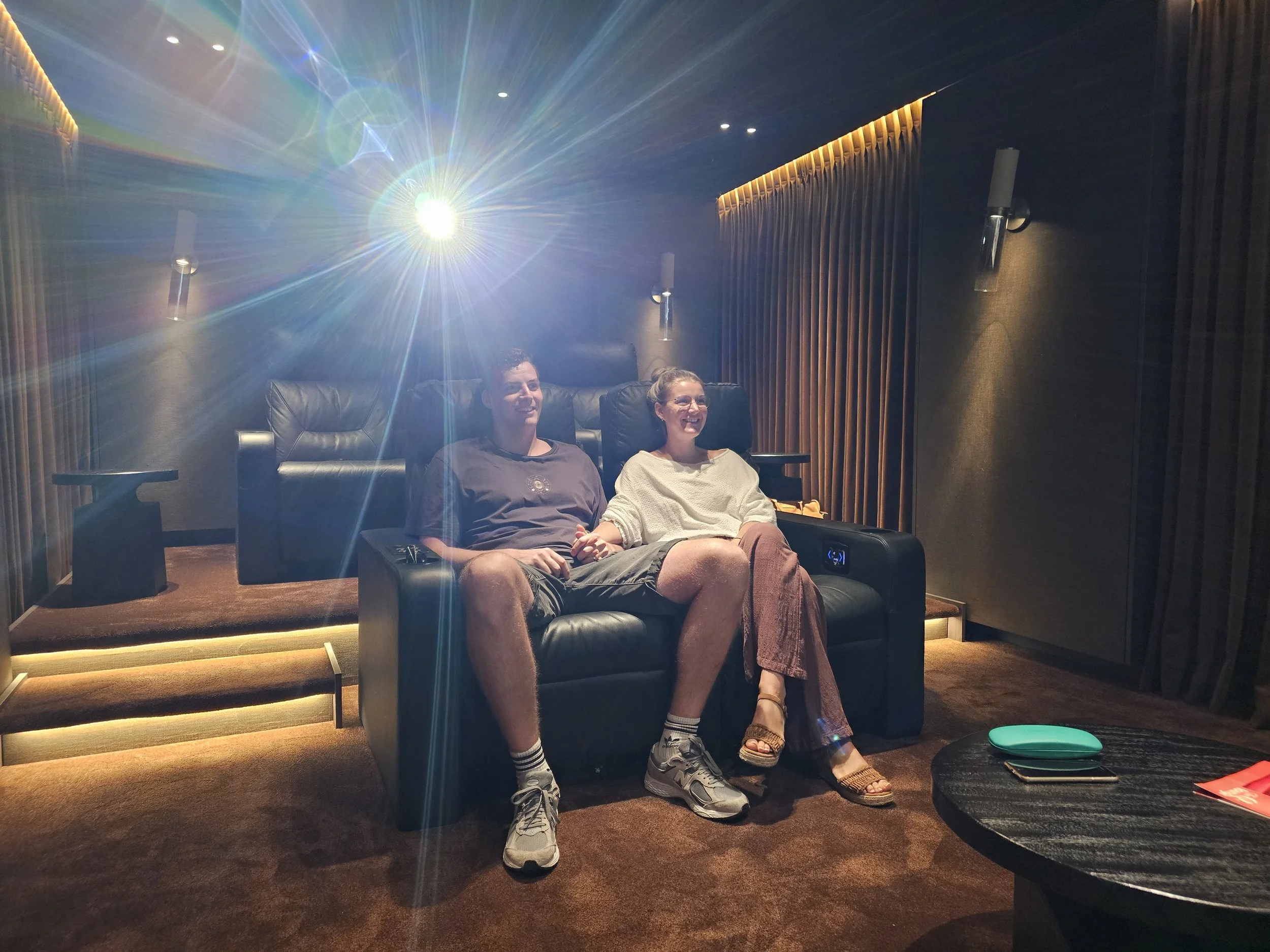 Twee mensen zitten ontspannen in een bioscoopzaal met zwart leren stoelen. Ze lachen en kijken naar het scherm. Er is een blauwe en groene glow op de vloer en er liggen een paar boeken of tijdschriften op een tafeltje voor hen.