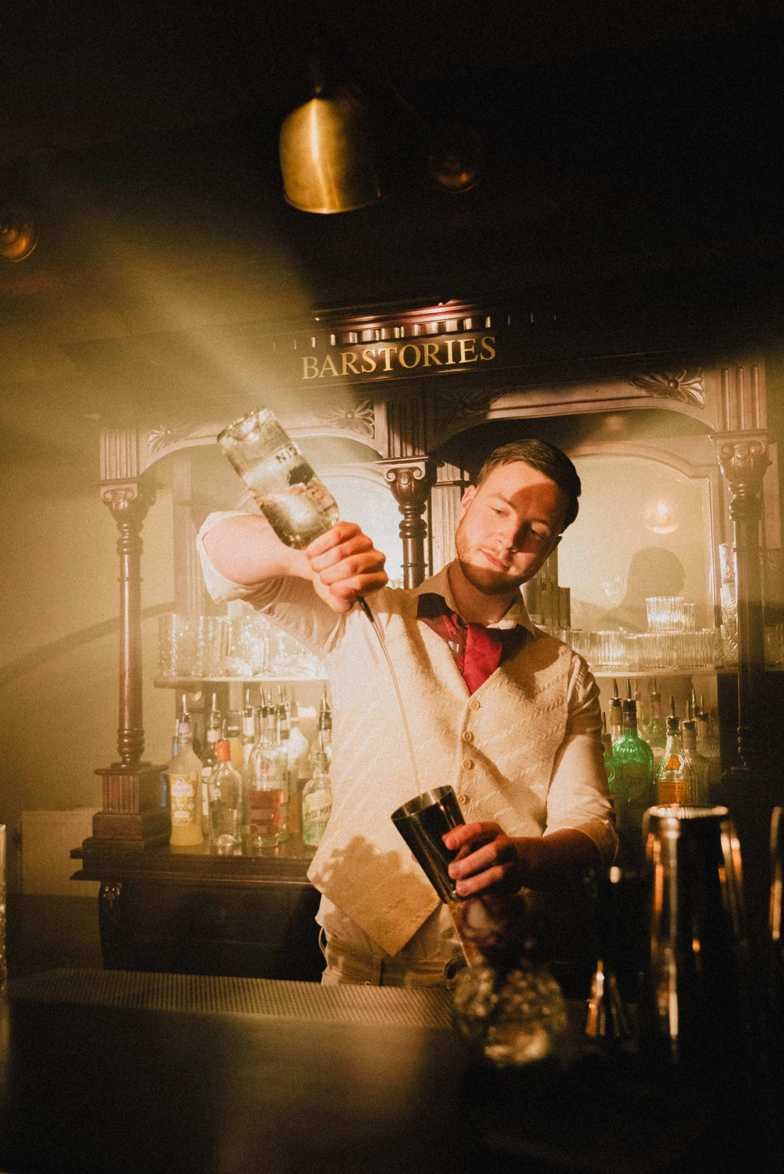 Man achter de bar die een cocktailshuim schept, met diverse flessen alcohol en specerijen op de voorgrond en een houten bar met spiegels op de achtergrond.