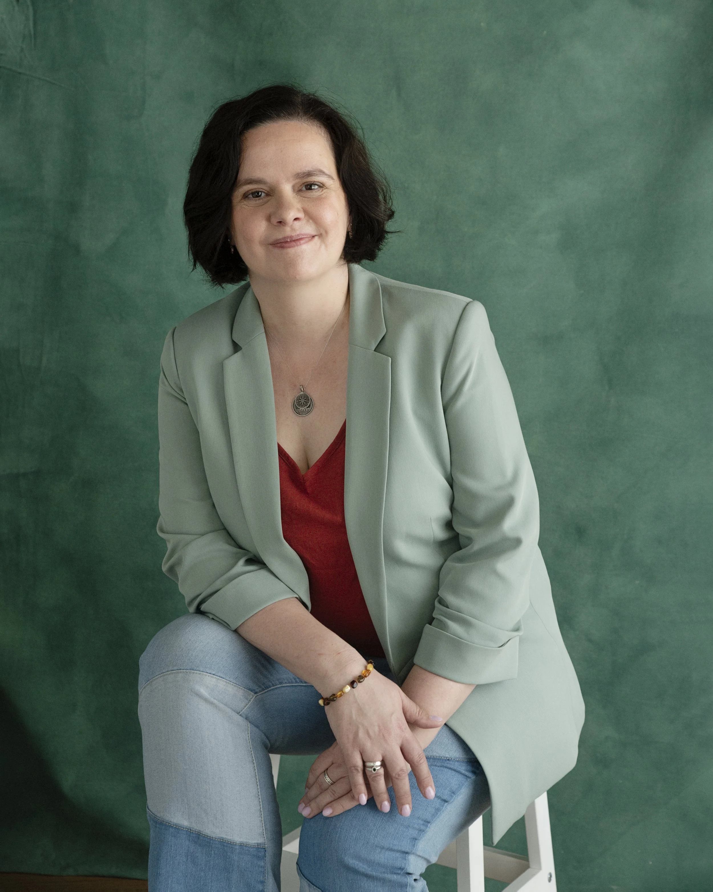 A woman with dark, curly, shoulder-length hair smiling at the camera. She is wearing a light green blazer over a red top, light blue jeans, and jewelry including a necklace, rings, earrings, and a multi-colored beaded bracelet. She is seated against a green background.