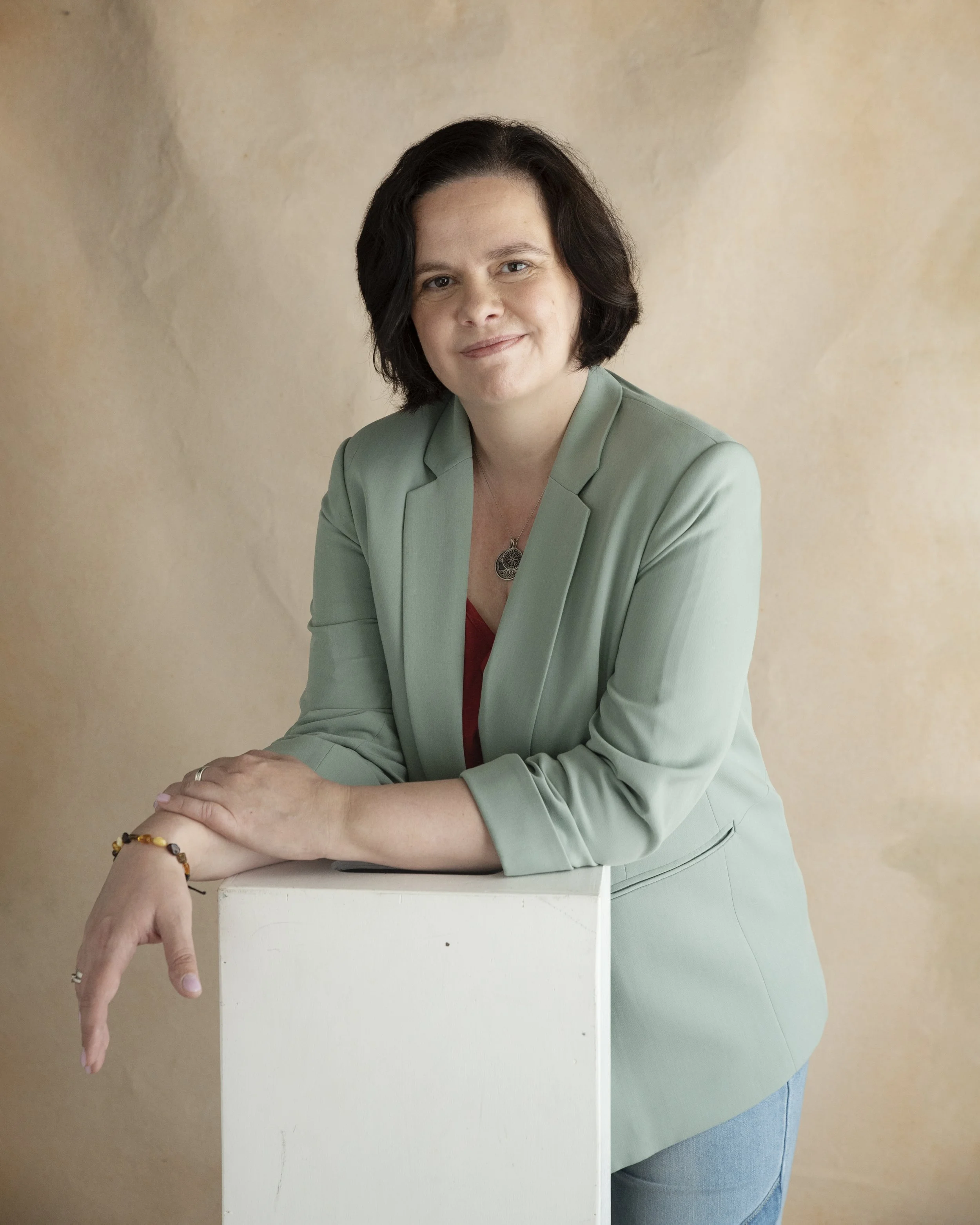 A woman with dark, shoulder-length hair wearing a green blazer and red shirt. She is smiling and has a necklace with a round pendant. She is leaning on a white podium, with her right hand resting on it, and wearing bracelets on her left wrist. The background is a plain beige wall.