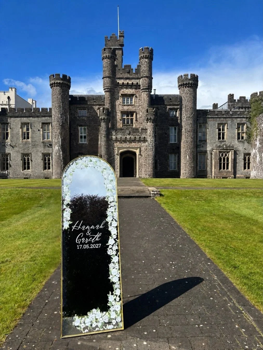 A castle with stone walls, towers, and battlements. In the foreground, a mirror with a decorative floral border and wedding date for Hannah and Gareth on May 17, 2027, standing on a paved walkway with green grass on either side.