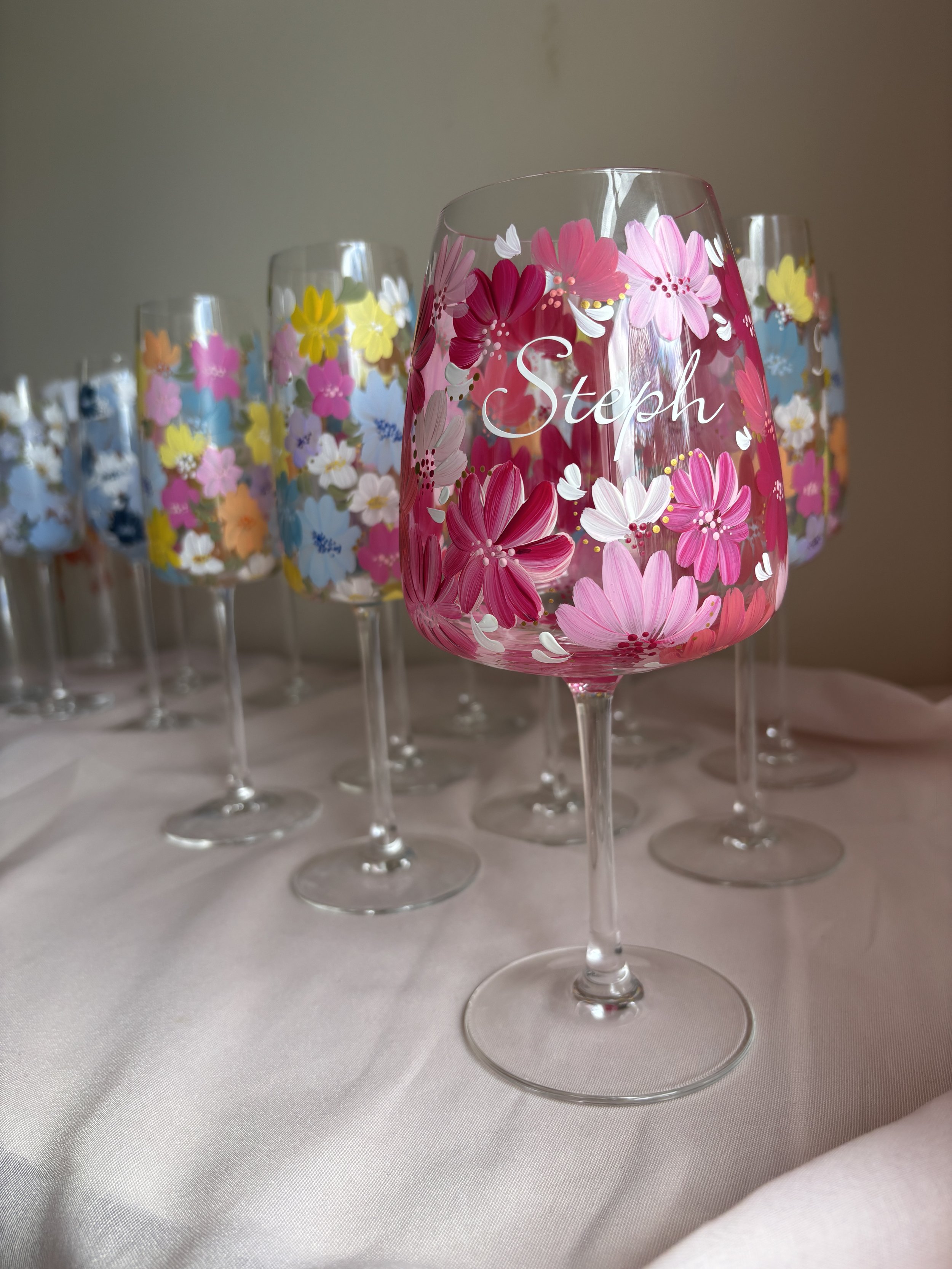 A set of five wine glasses with colorful floral designs, with one glass in the foreground featuring the name 'Steph' written on it.