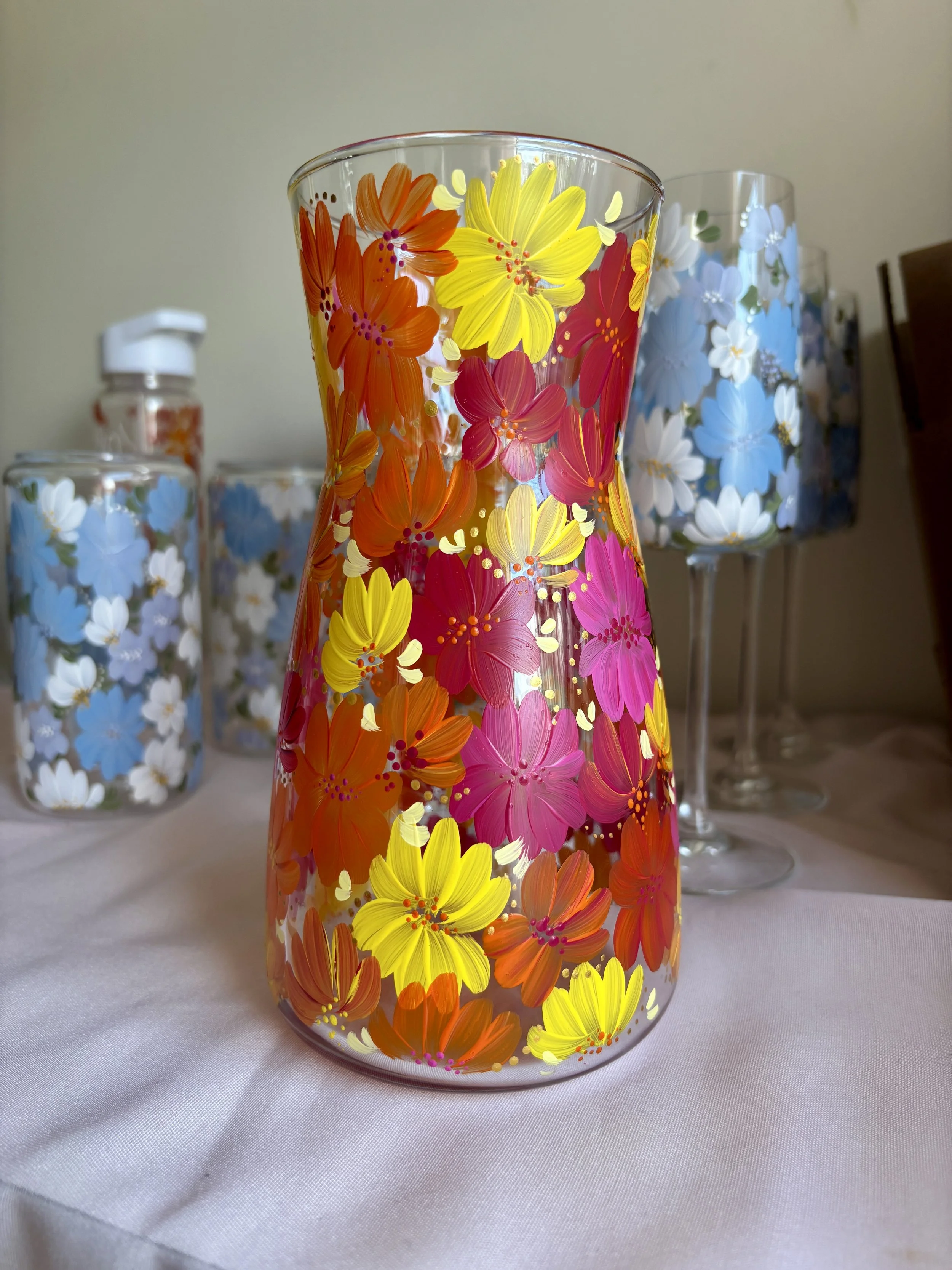 A clear glass vase decorated with yellow, pink, and orange flowers. In the background are two matching glasses with white and blue flowers.