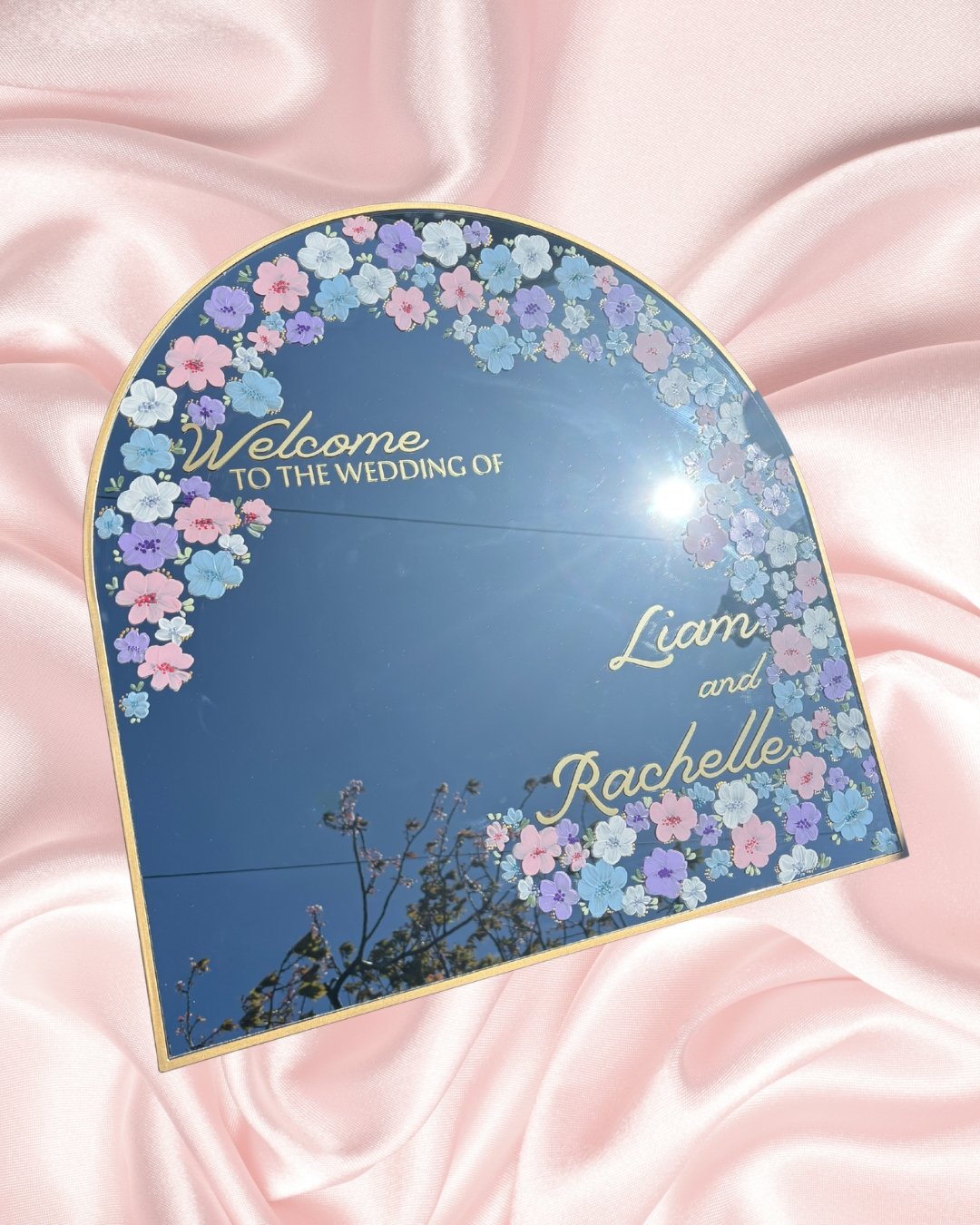 Wedding welcome mirror with pink, purple, and blue flowers border, reading 'Welcome to the wedding of Liam and Rachelle,' set on pink satin fabric.