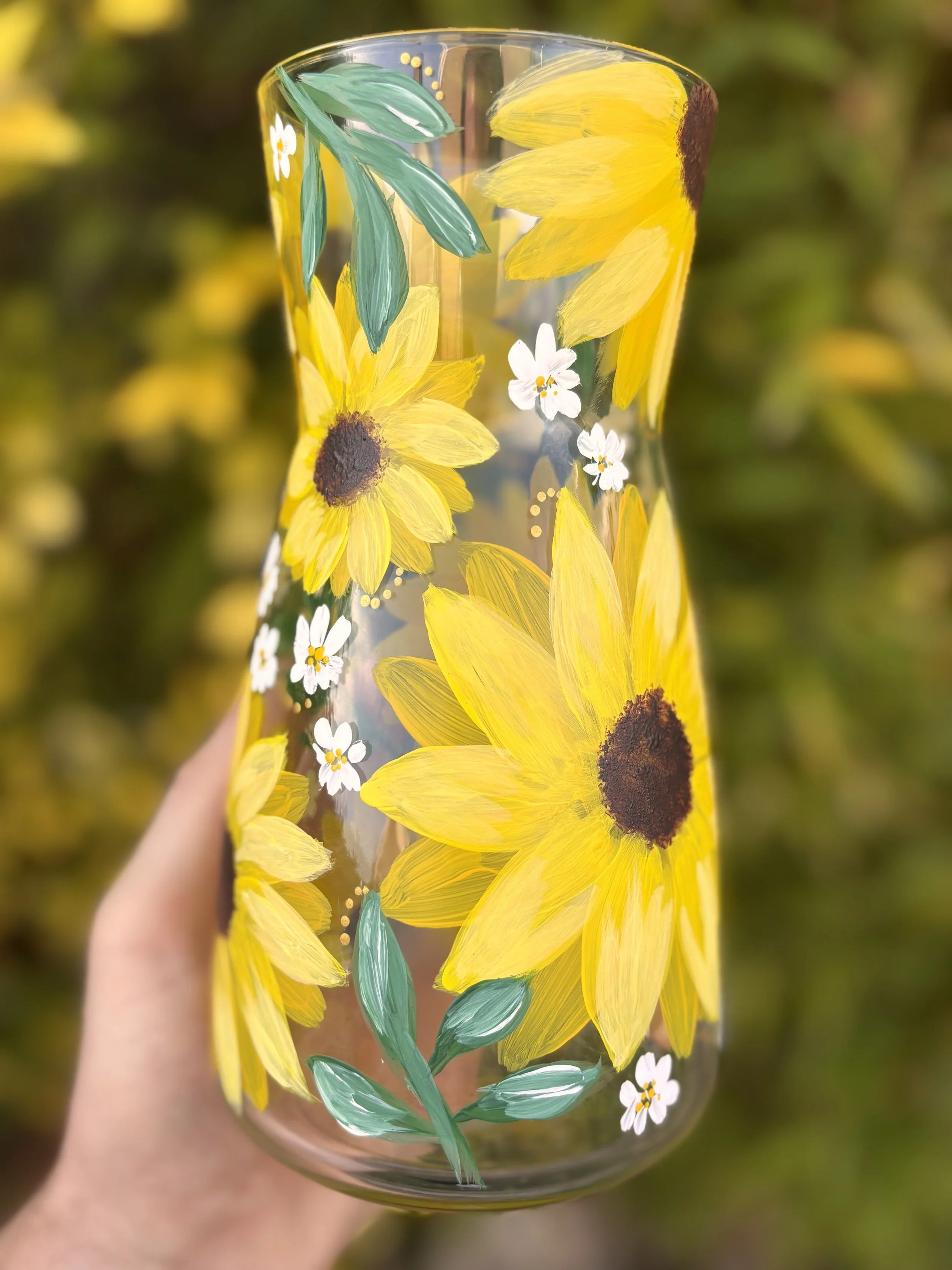A hand holding a glass vase with a painted sunflower design, featuring yellow petals, green leaves, and small white flowers, against a blurred outdoor background.