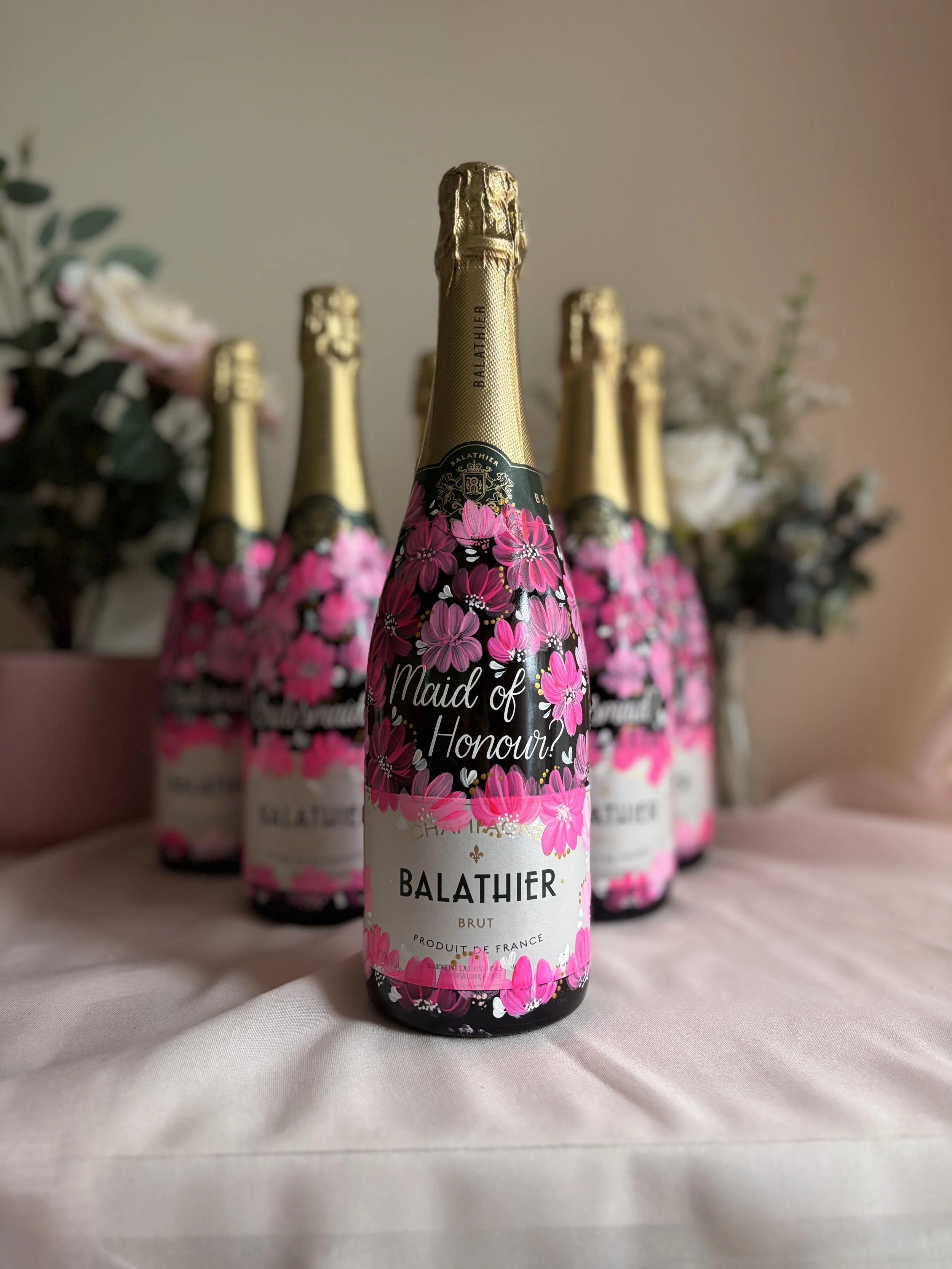 Several bottles of prosecco with painted pink floral designs, and white text with Bridesmaid and Maid of Honour on the bottles.