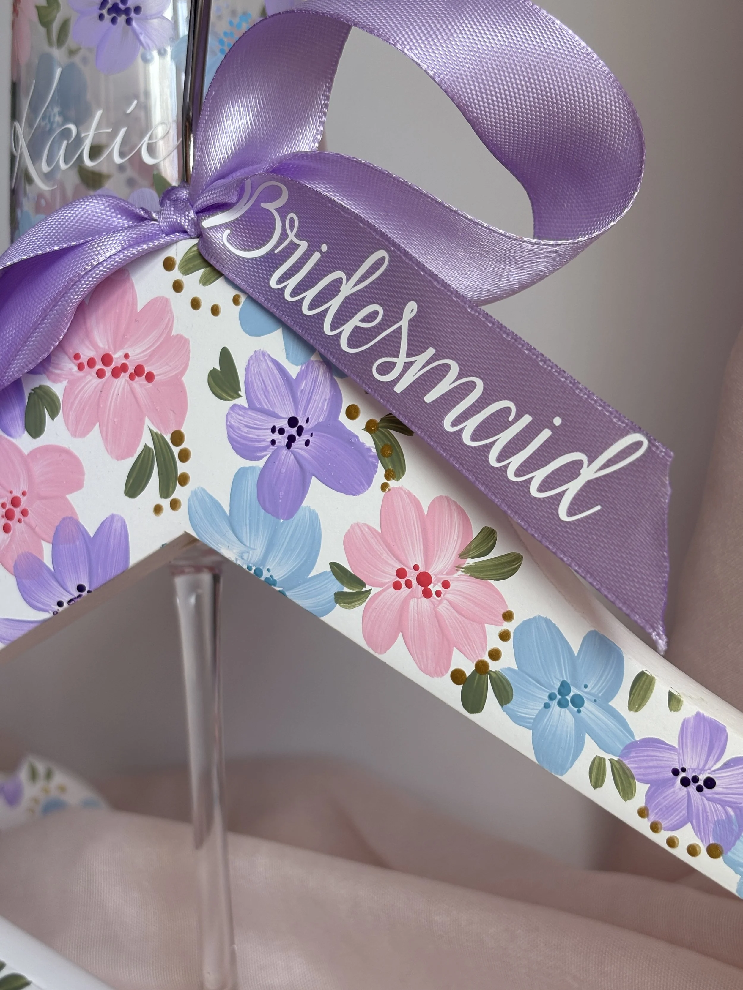 A coat hanger decorated with painted pink, purple, and blue flowers, with a purple satin ribbon that says 'Bridesmaid.'