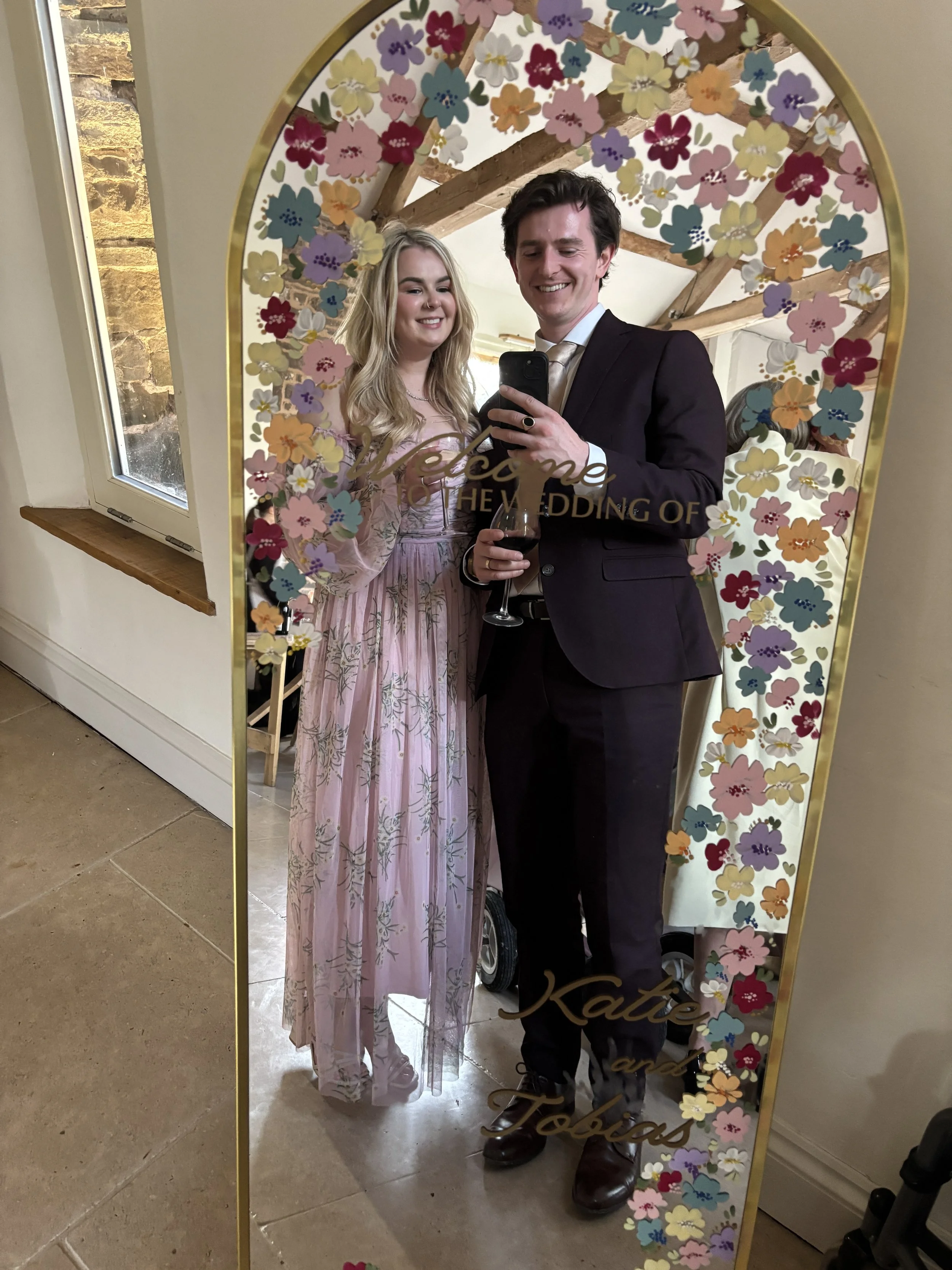 A young woman and a man taking a selfie in a mirror with floral decoration and the words 'Welcome to the wedding of' written on the mirror. The woman is wearing a pink dress, and the man is dressed in a suit and tie, holding a glass of red wine.