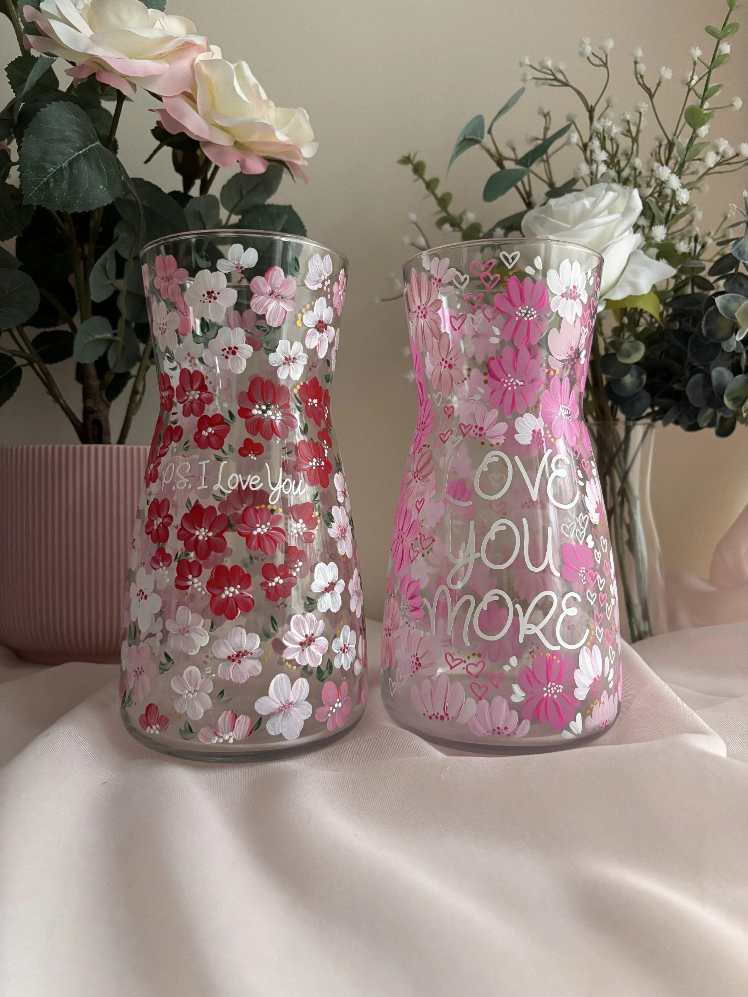 Two glass vases with pink and white floral designs, one with the words "P.S. I Love You" and the other with "Love You More", are set on a white fabric surface with a backdrop of pink and white artificial flowers and greenery.