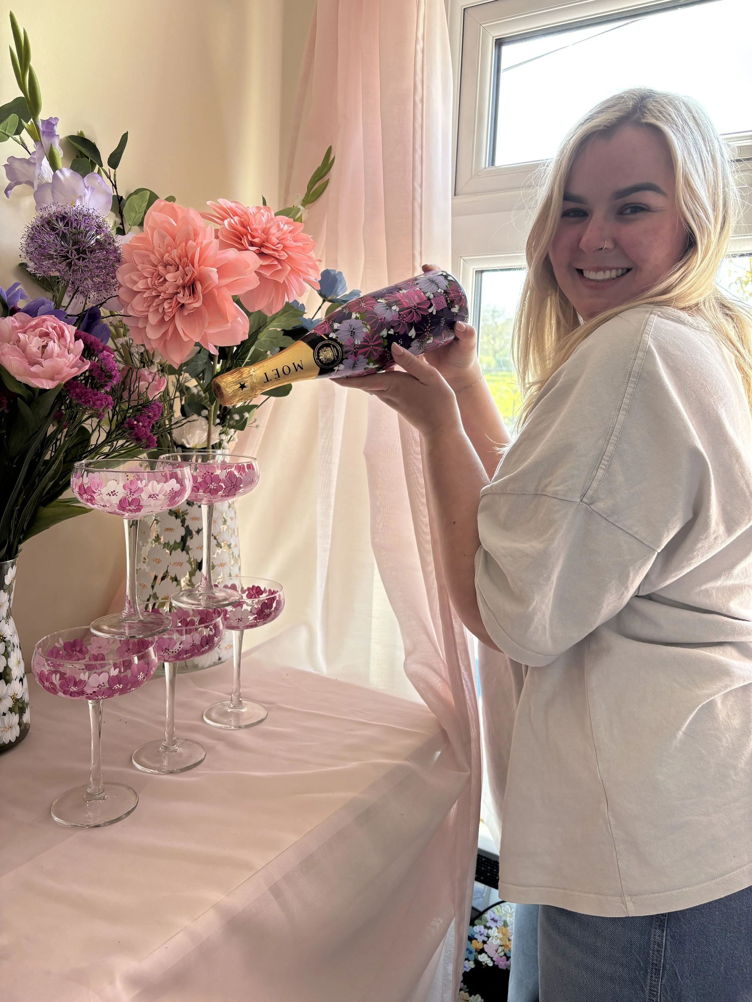 A woman with blonde hair pretends to pour a bottle of champagne into a champagne glass on top of a pyramid of glasses.
