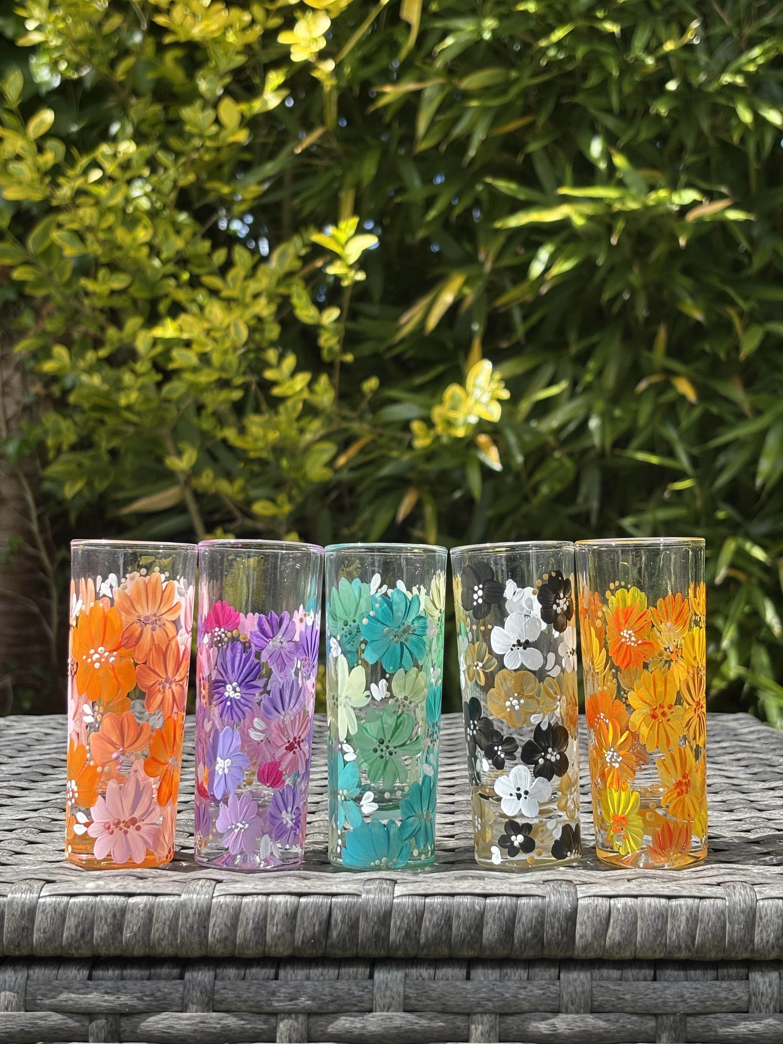 Six colourful shot glasses decorated with floral patterns are arranged in a row on a woven surface, with greenery in the background.