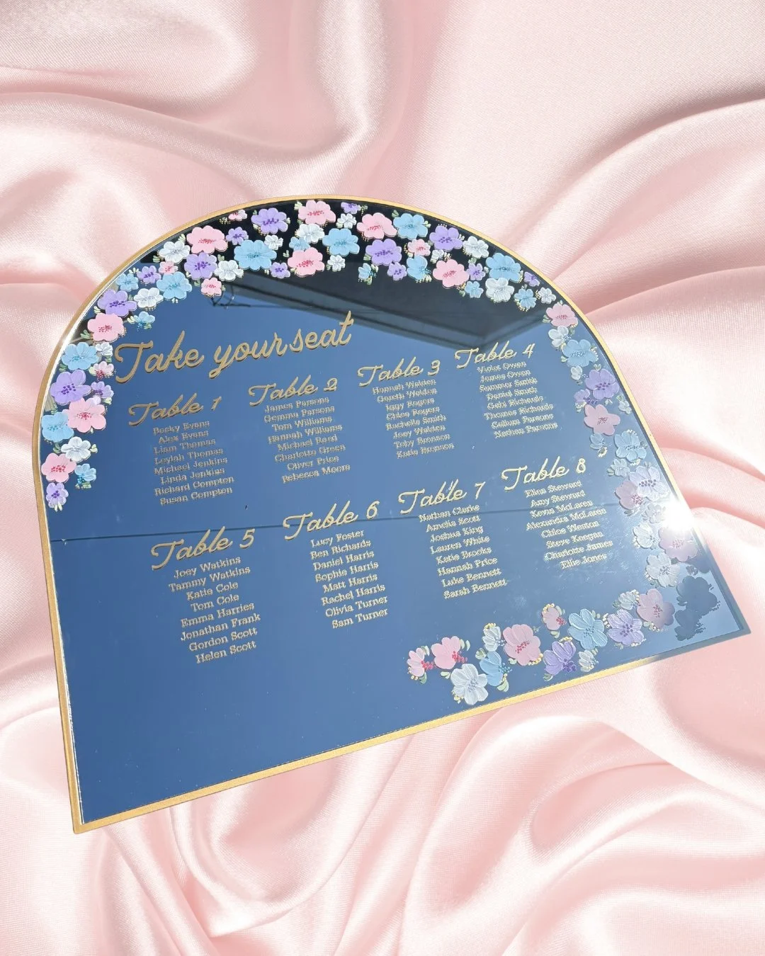 Mirrored seating chart with flower border and gold accents, titled 'Take your seat', listing names by table number from 1 to 8, on pink satin background.