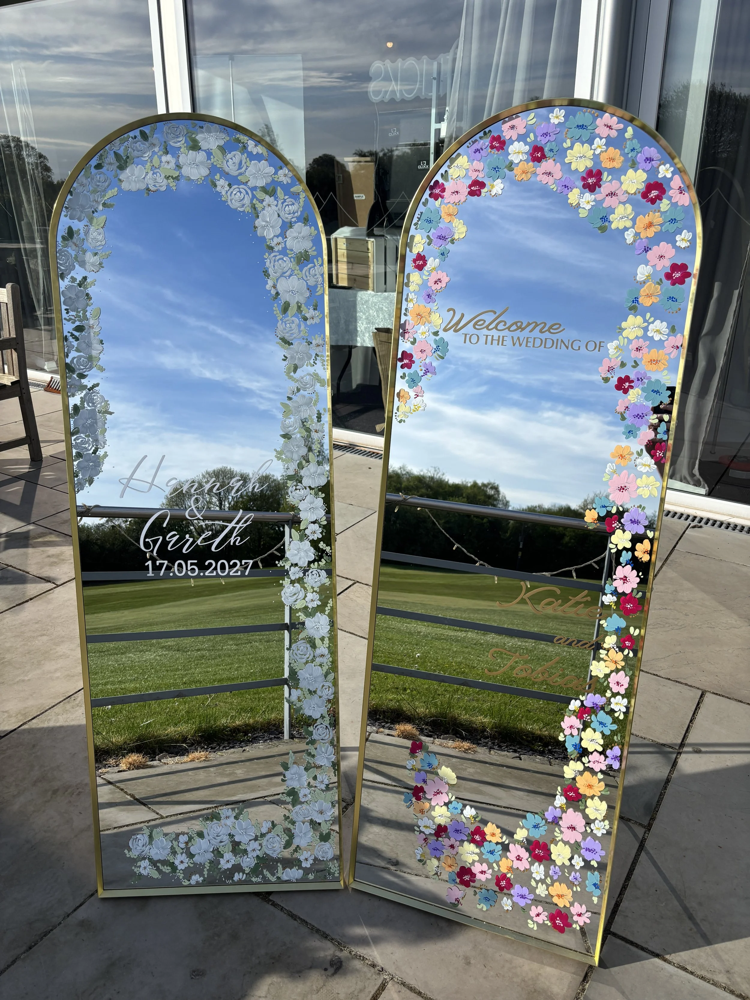 Two decorative mirrors, full length and upright, with painted flowers around the borders.  The flowers are in white on one mirror and pastel colours on the other. There's gold text on them welcoming guests to the weddings.