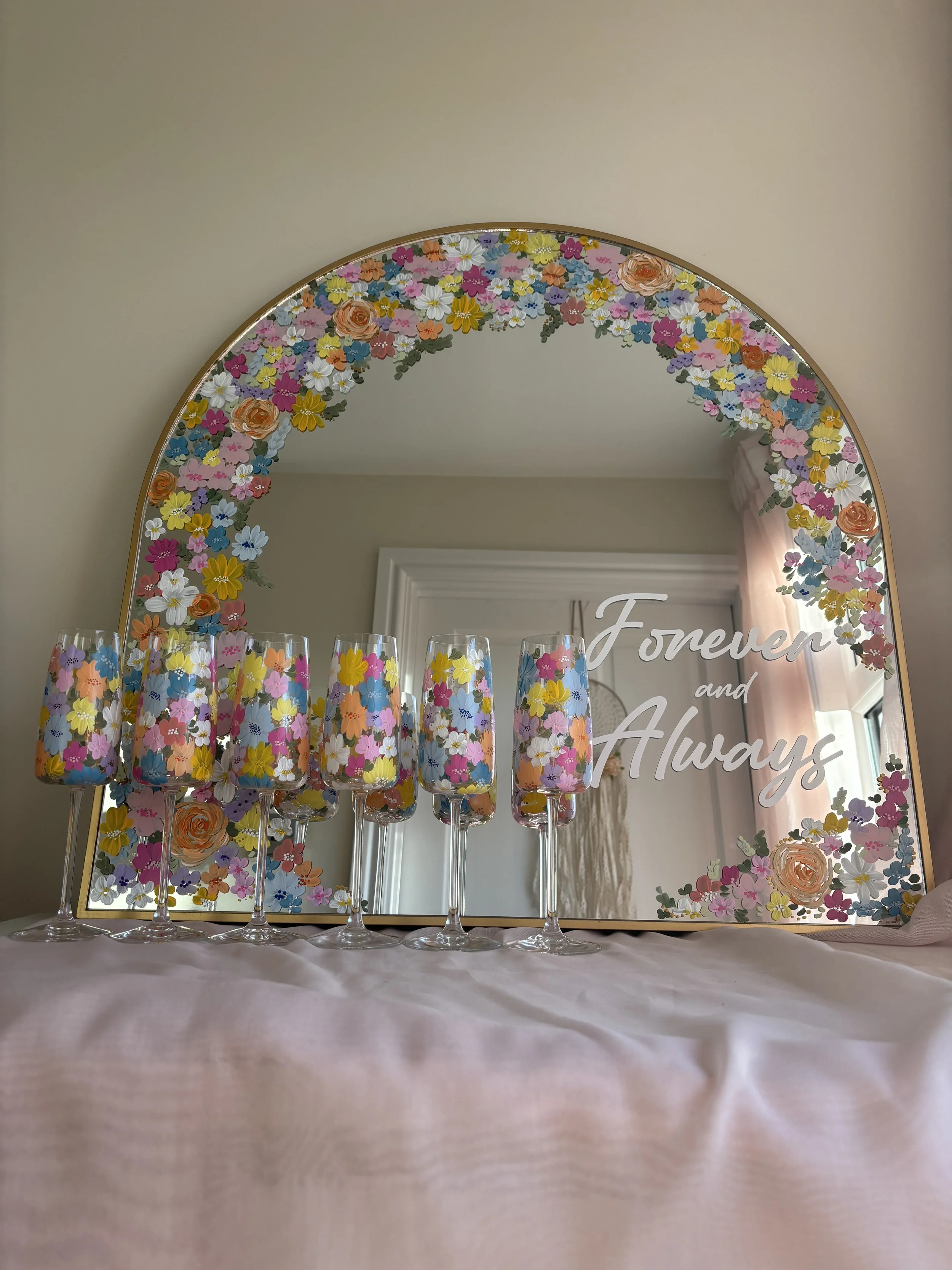 Decorative mirror with a floral border, the words "Forever and Always" on the mirror, and multiple colourful flute glasses with floral patterns on a table.