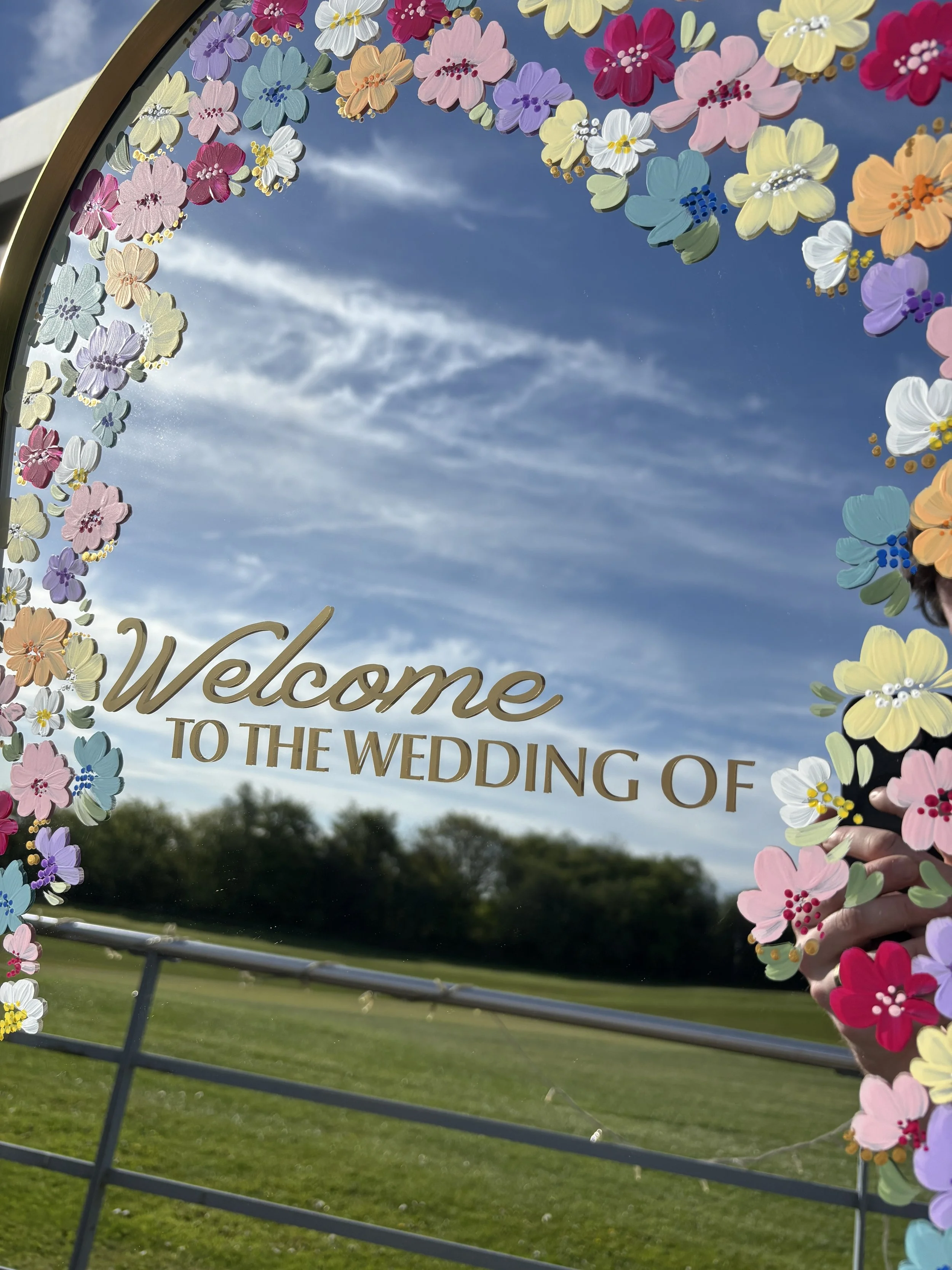 A decorative wedding mirror framed with colorful painted flowers in pastel colours like pink, purple, yellow, and white, with a background of a blue sky with scattered clouds, green grass, and trees.