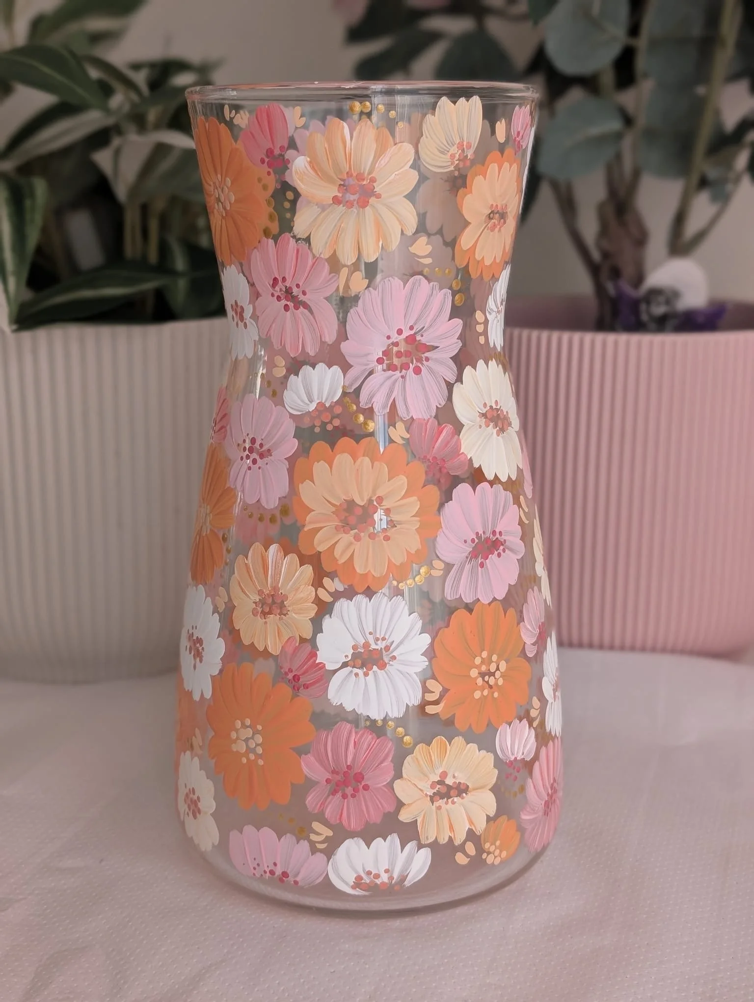 A clear glass vase with a floral pattern featuring pink, peach, white, and orange flowers, placed on a light surface with leafy green plants.