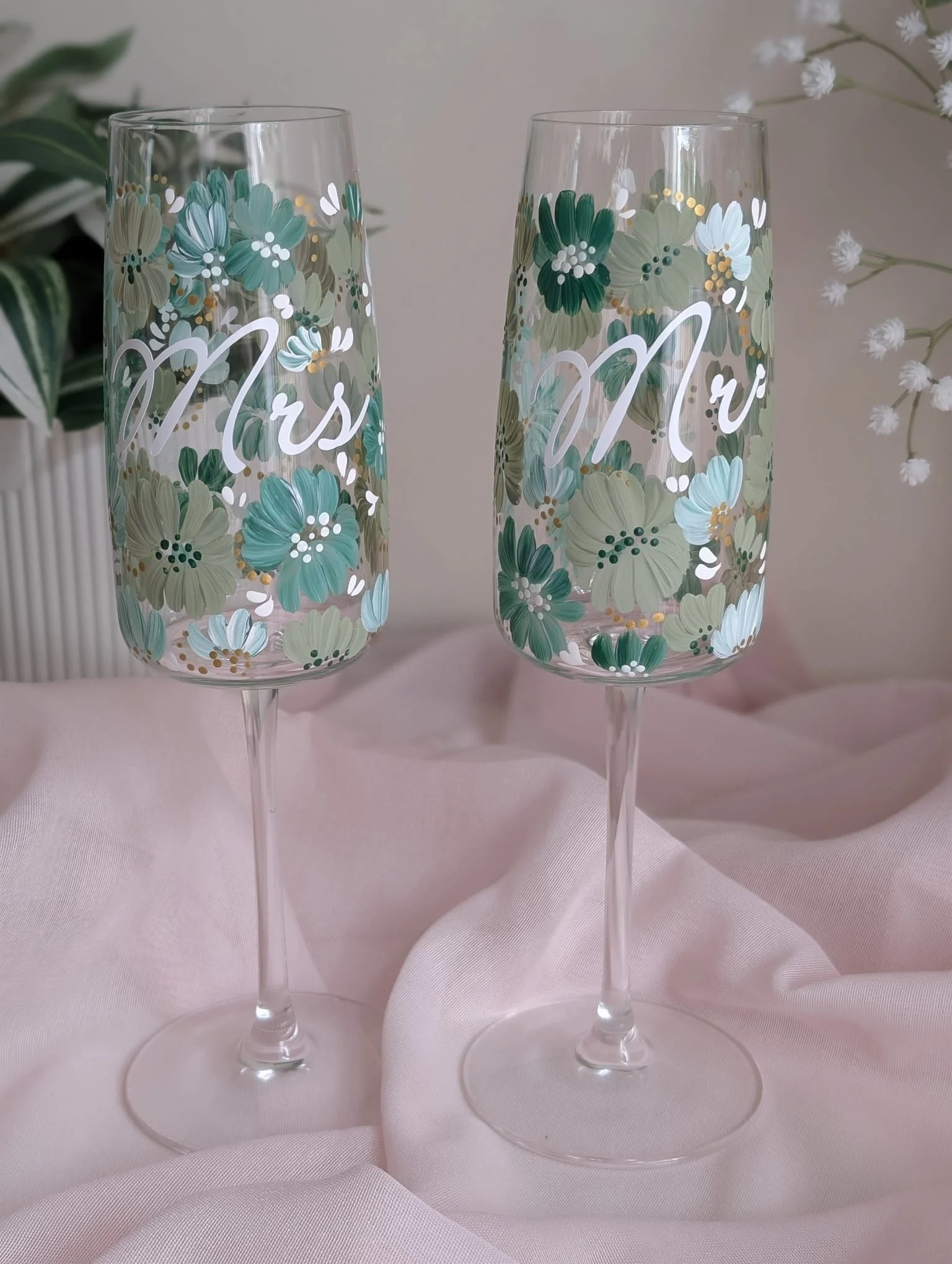 Two tall champagne flutes decorated with floral patterns and the words "Mrs" and "Mr" written in white, set on a pale pink tablecloth with flowers in the background.