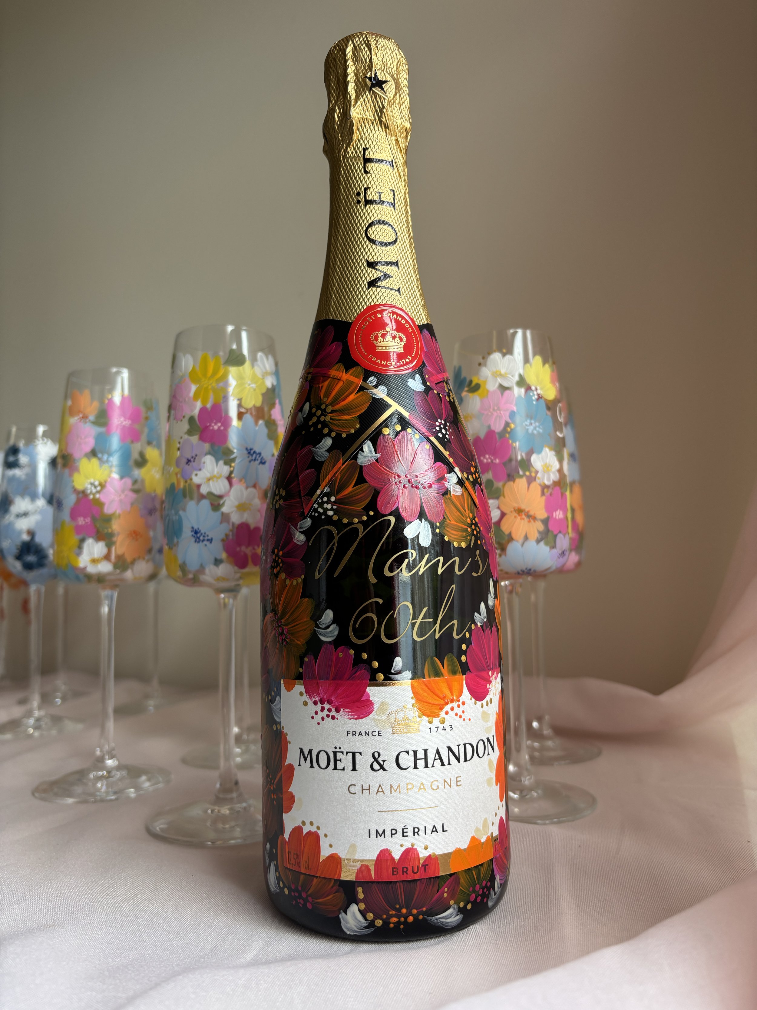 A decorated bottle of Moët & Chandon champagne with floral artwork, surrounded by four champagne flutes with matching floral designs, set on a white cloth surface.