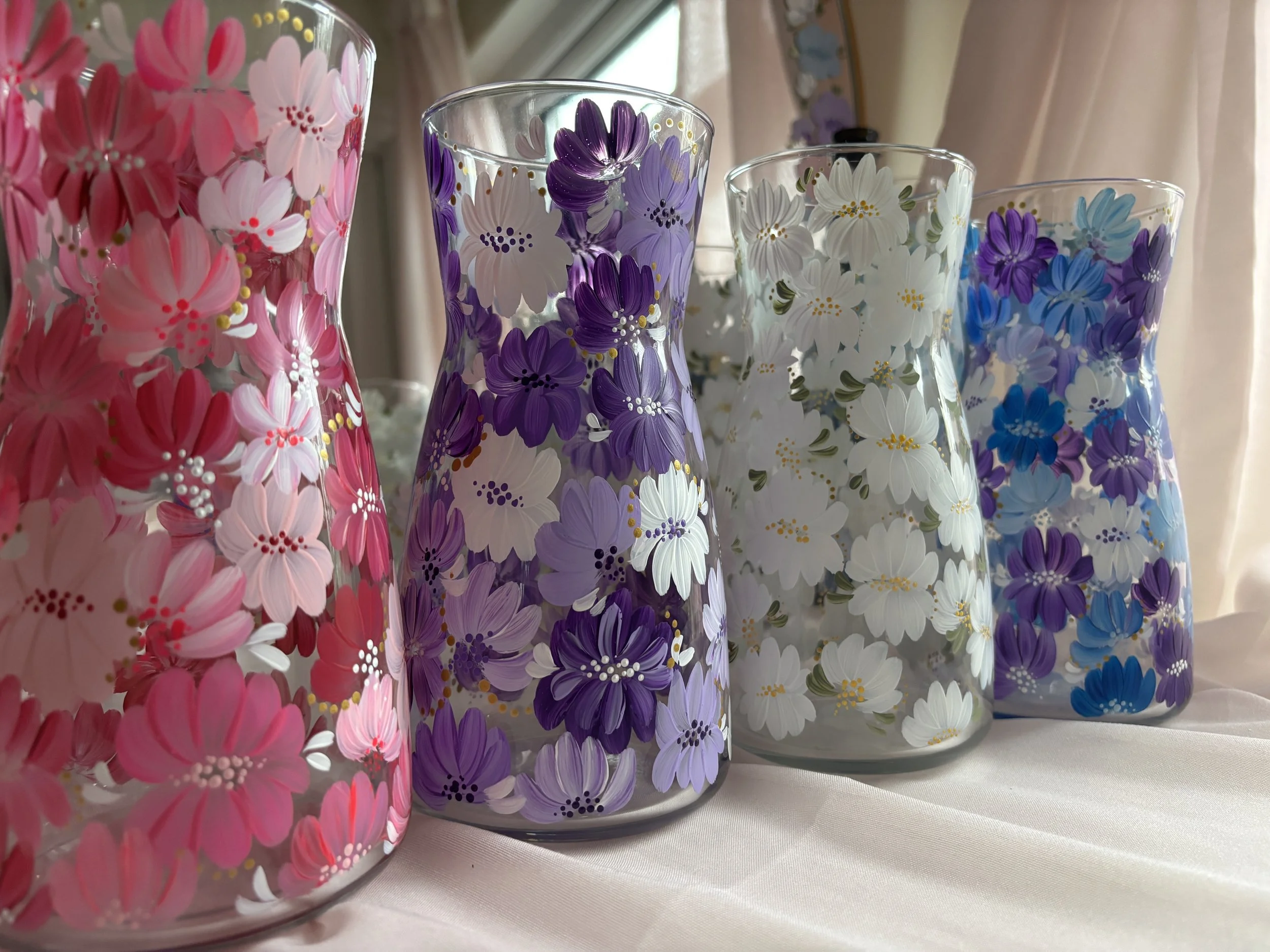 Close-up of four clear glass vases with hand painted flowers. The vases contain painted flowers in shades of pink, purple, white, and blue, with some yellow accents.