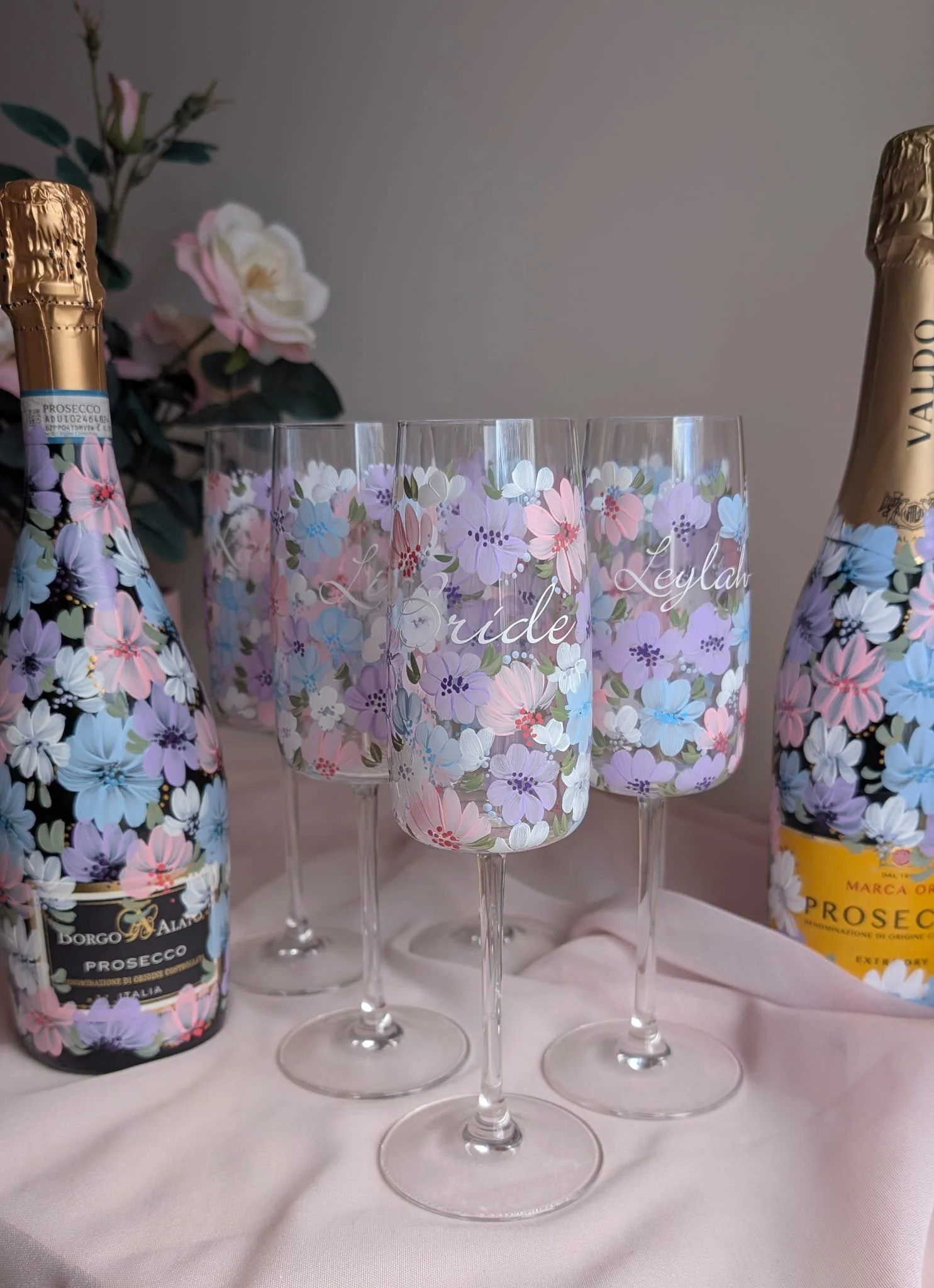 Four prosecco glasses with floral patterns and personalised writing, flanked by two bottles of prosecco, on a white tablecloth with pink and white flowers in the background.