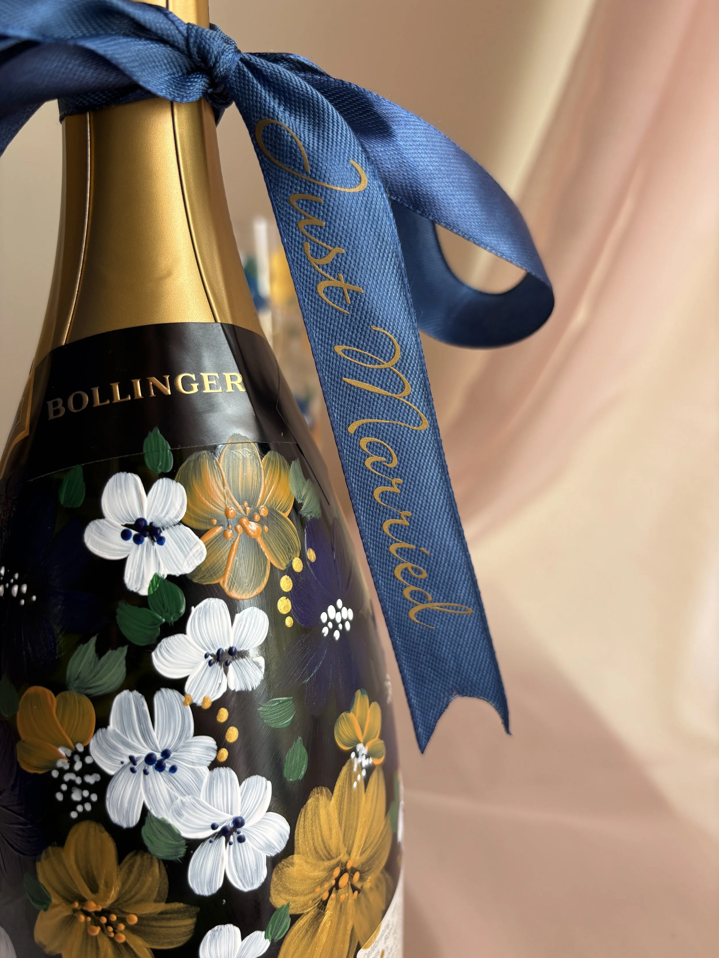 Close-up of a champagne bottle with a blue silk ribbon tied around its neck. The text on the ribbon reads "Just Married" and the bottle features a floral design with white and gold flowers.