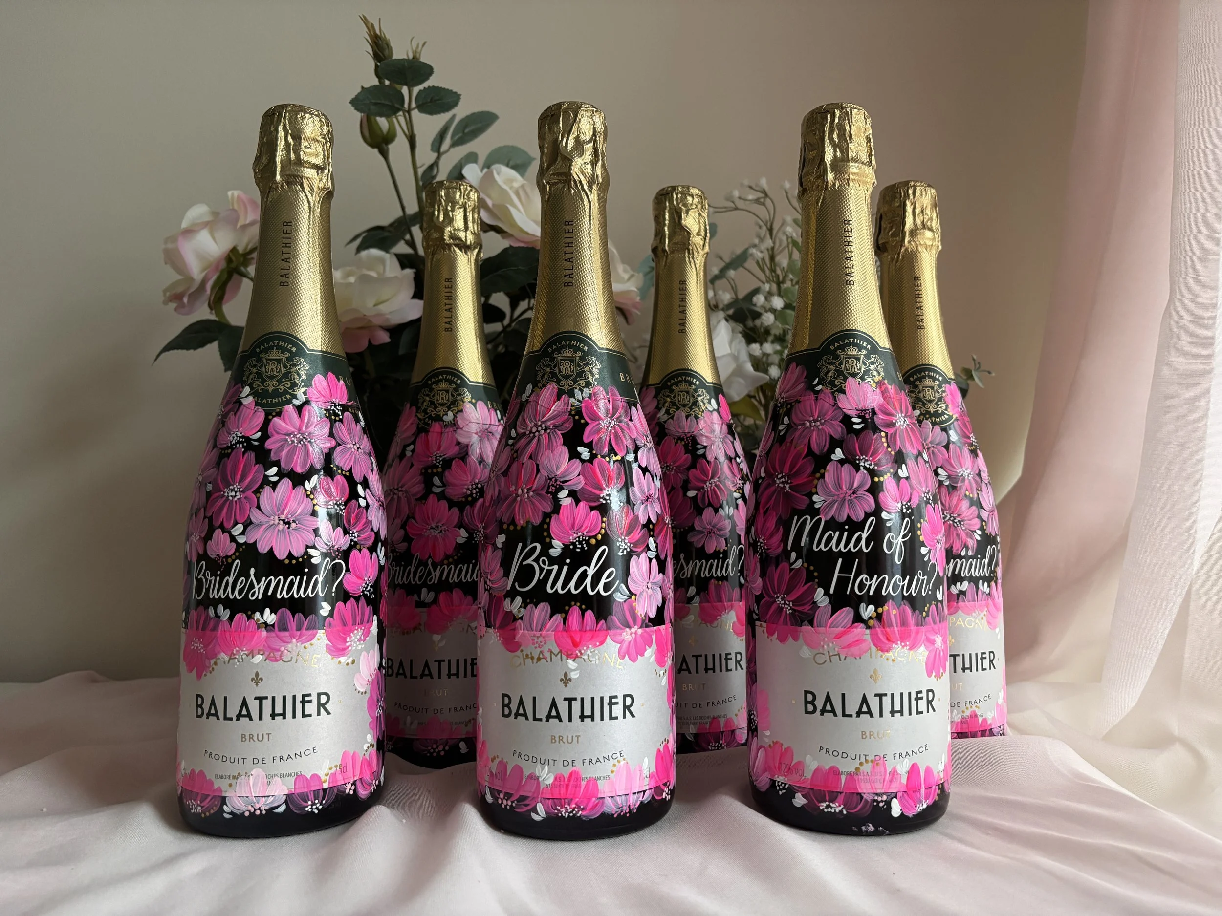 Six bottles of champagne with pink floral labels standing upright on a white surface, with pink and white flowers and a pink curtain in the background. There are titles on the bottles, like Bridesmaid and Maid of Honour, used for bridesmaid proposals