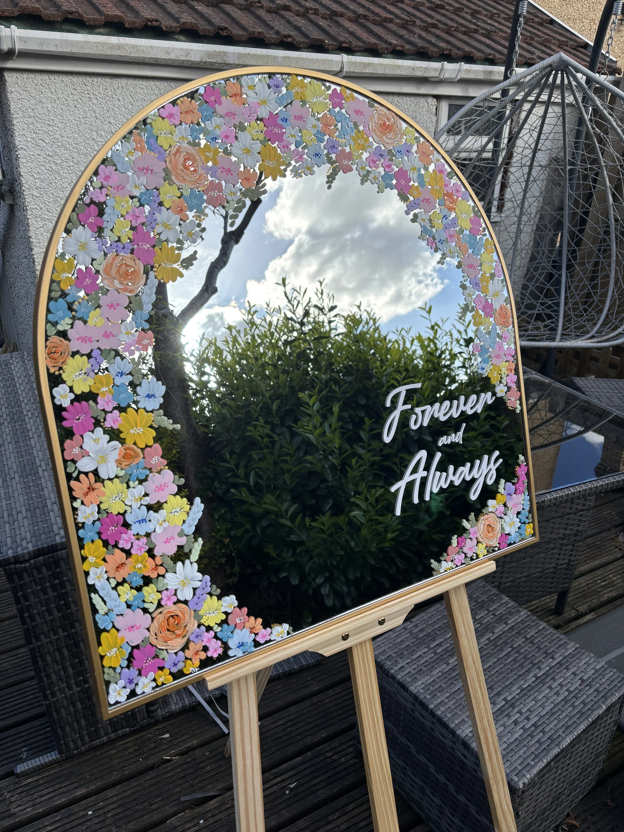 A decorative mirror with a floral border and the words "Forever and Always" written on it. The mirror reflects the sky, clouds, a tree, and some greenery. It is positioned on a wooden stand outdoors, next to wicker furniture.