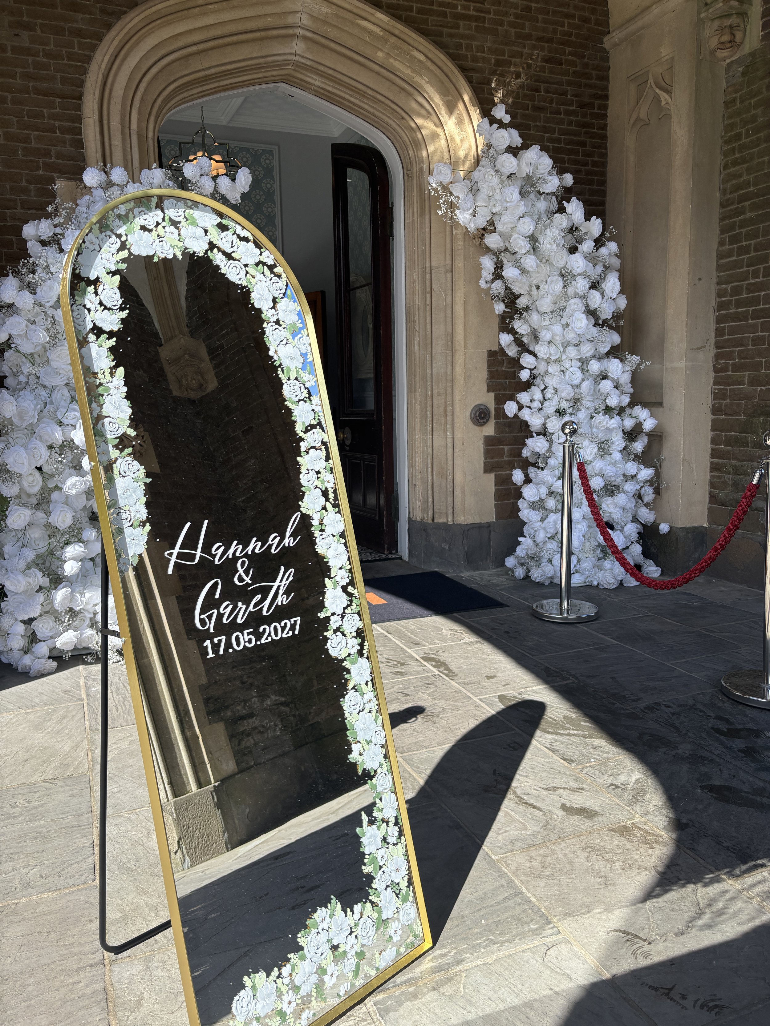 Wedding invitation sign next to an arch of white roses outside a building entrance. The sign has a black background with white text reading "Hannah & Gareth 17.05.2027" and is decorated with a floral border. The building has a brick facade with a dec