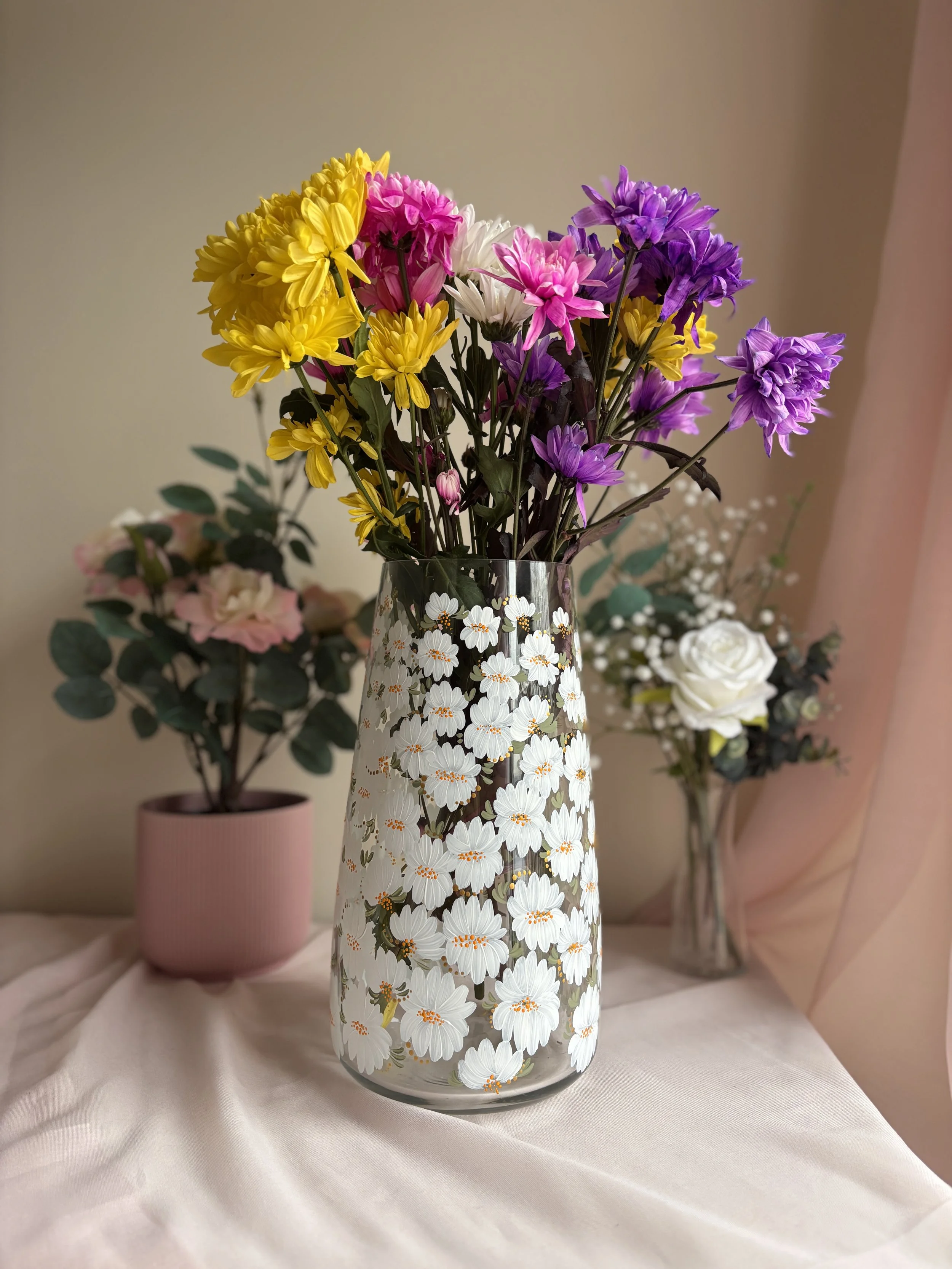 A large glass vase filled with colorful flowers, including yellow, pink, white, and purple blossoms, placed on a white fabric surface with a blurred background of additional flowers and greenery. The vase is painted with white flowers.