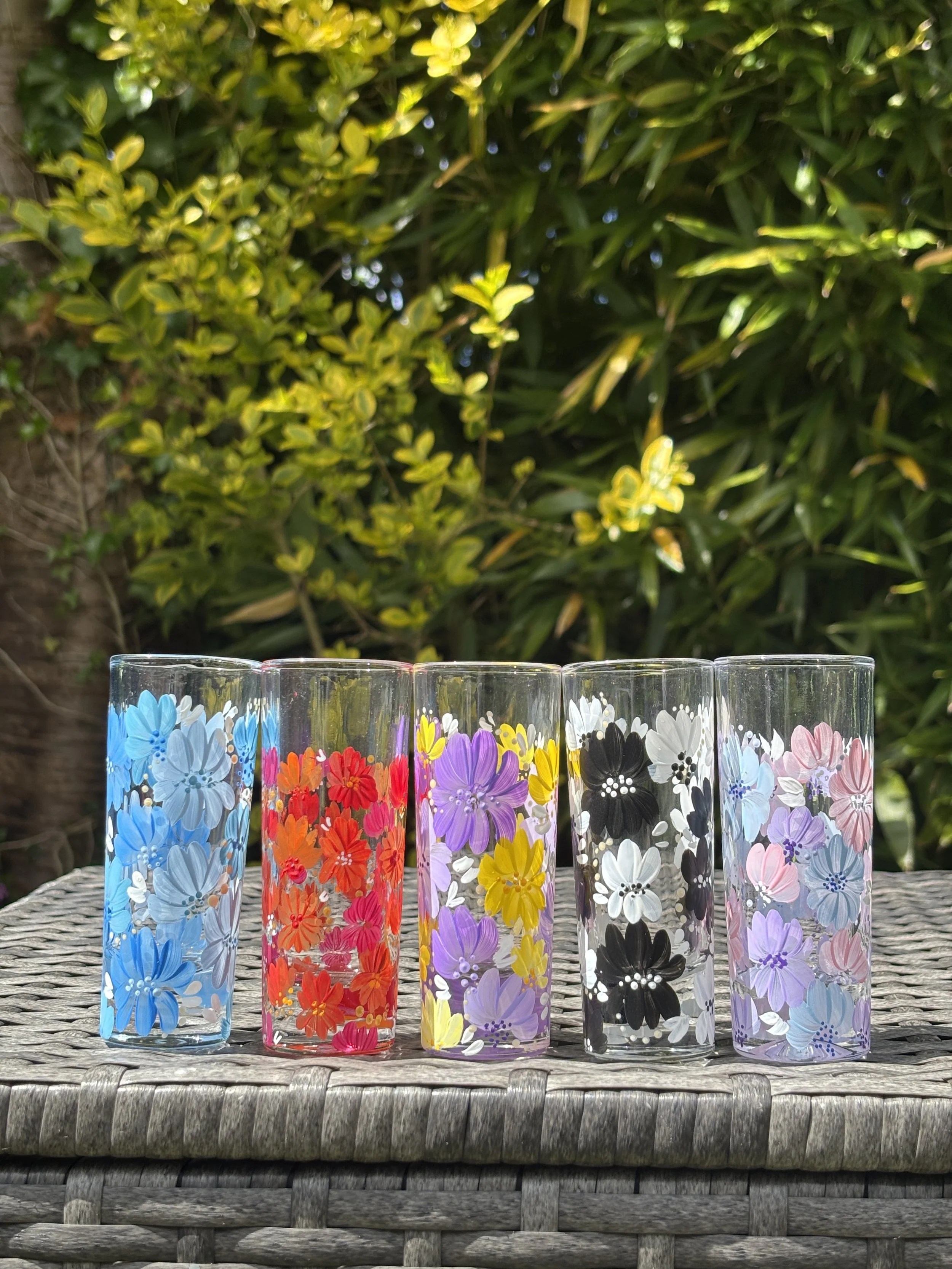 Six colourful shot glasses with floral patterns, arranged in a row on a textured wicker surface outdoors, with leafy green bushes in the background.