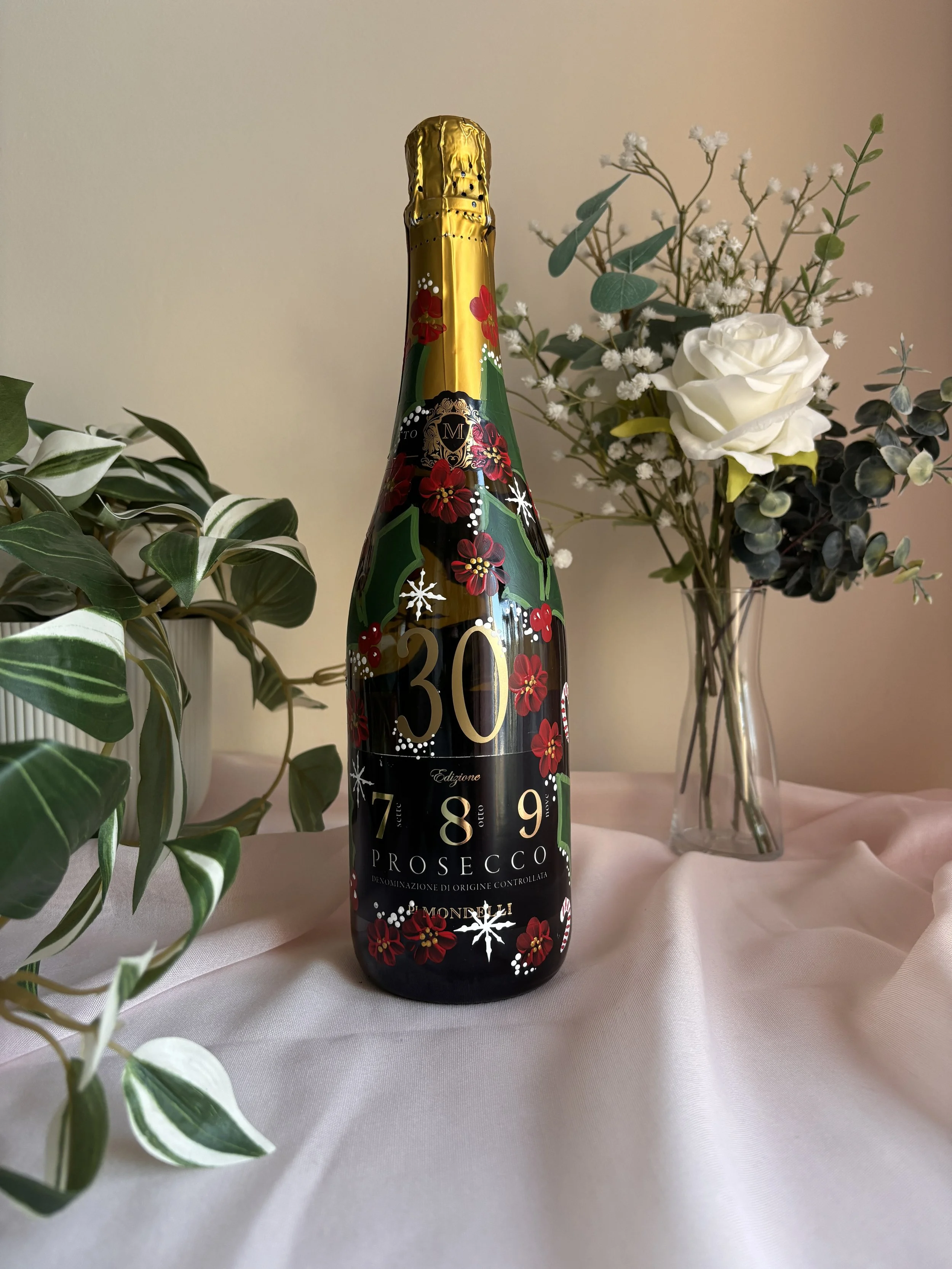 A bottle of Prosecco with Christmas-themed decorations, placed on a white surface between a green leafy plant on the left and a glass vase with white flowers and green foliage on the right.