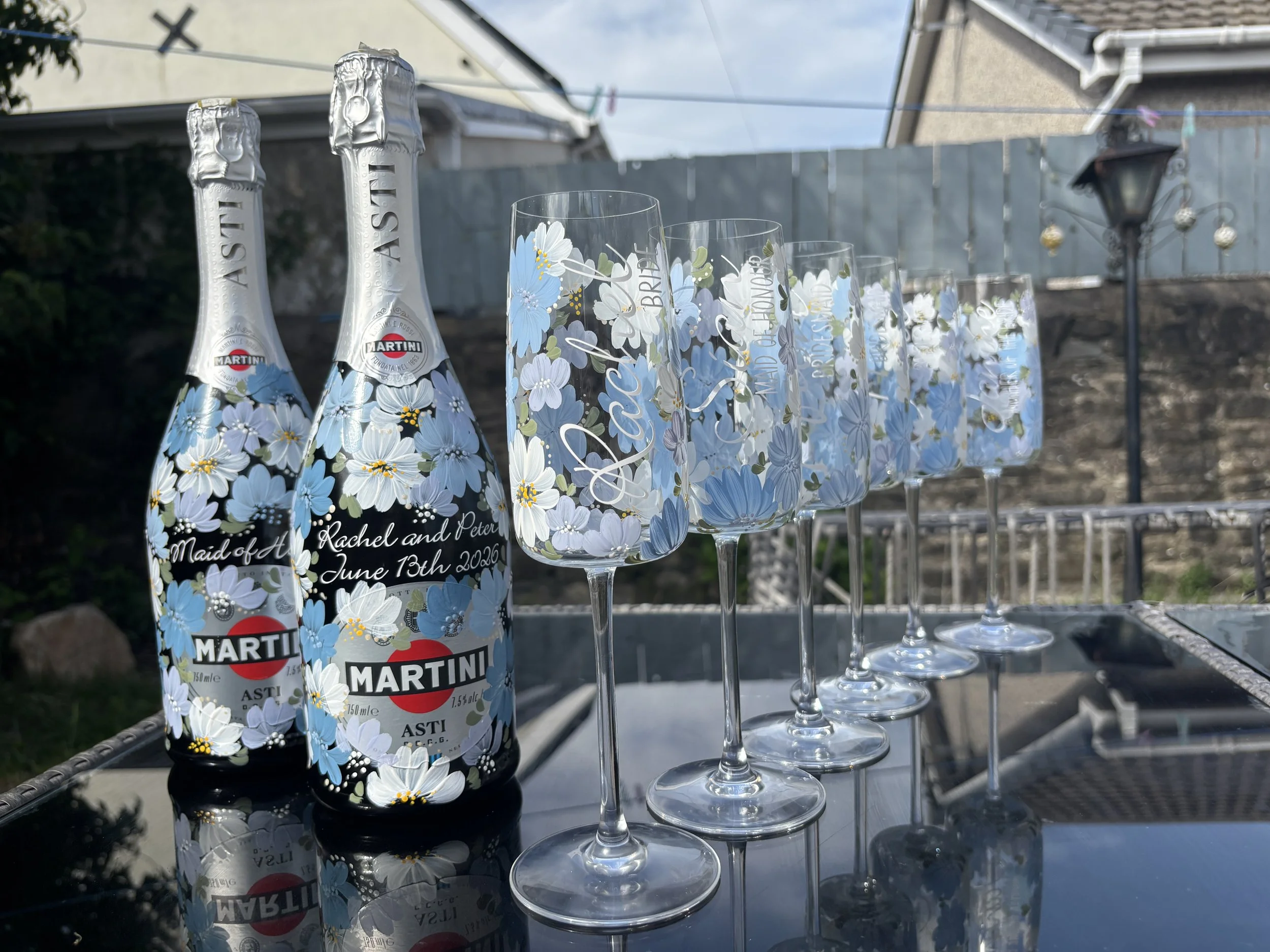 Two bottles of Martini Asti sparkling wine with blue and white floral designs painted on them, placed on a reflective surface outdoors, next to several champagne glasses with similar floral design.